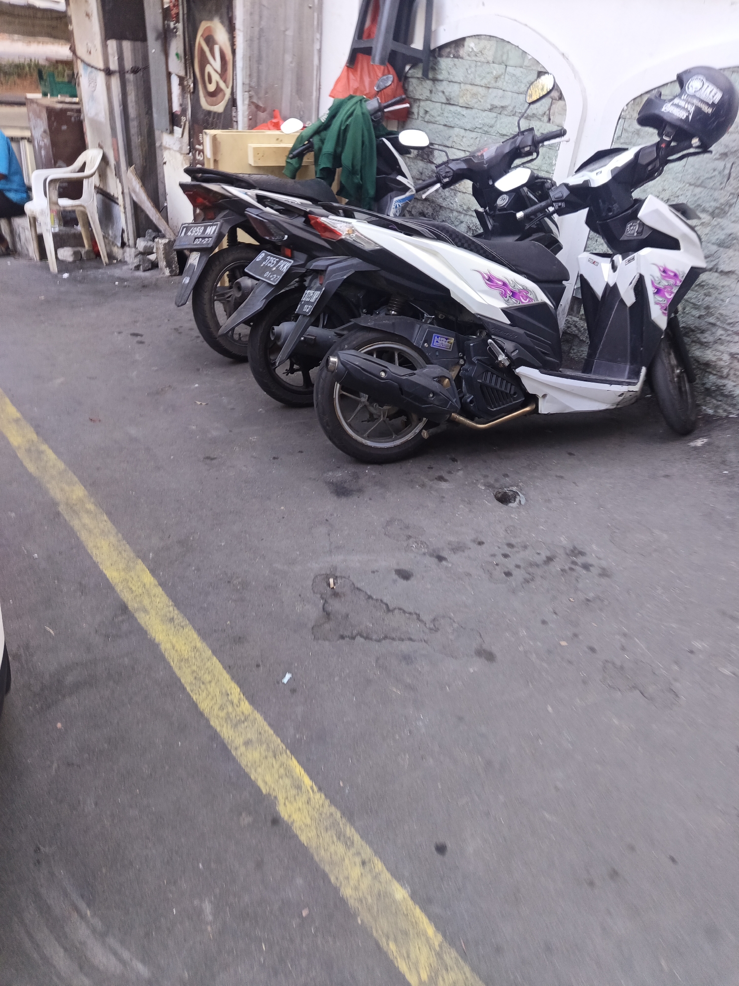 Two automatic scooters are parked on an asphalt surface marked with a yellow line. The scooter in the foreground is white and black with purple graphics on the side panels. It has a black helmet resting on its handlebars. The scooter behind it is mostly black. A white plastic chair is visible to the left of the scooters, near a brick wall and a doorway. The scene appears to be in an urban setting, likely a street or alleyway. The presence of the scooters and the general urban decay suggests this location is in North Jakarta, Indonesia.