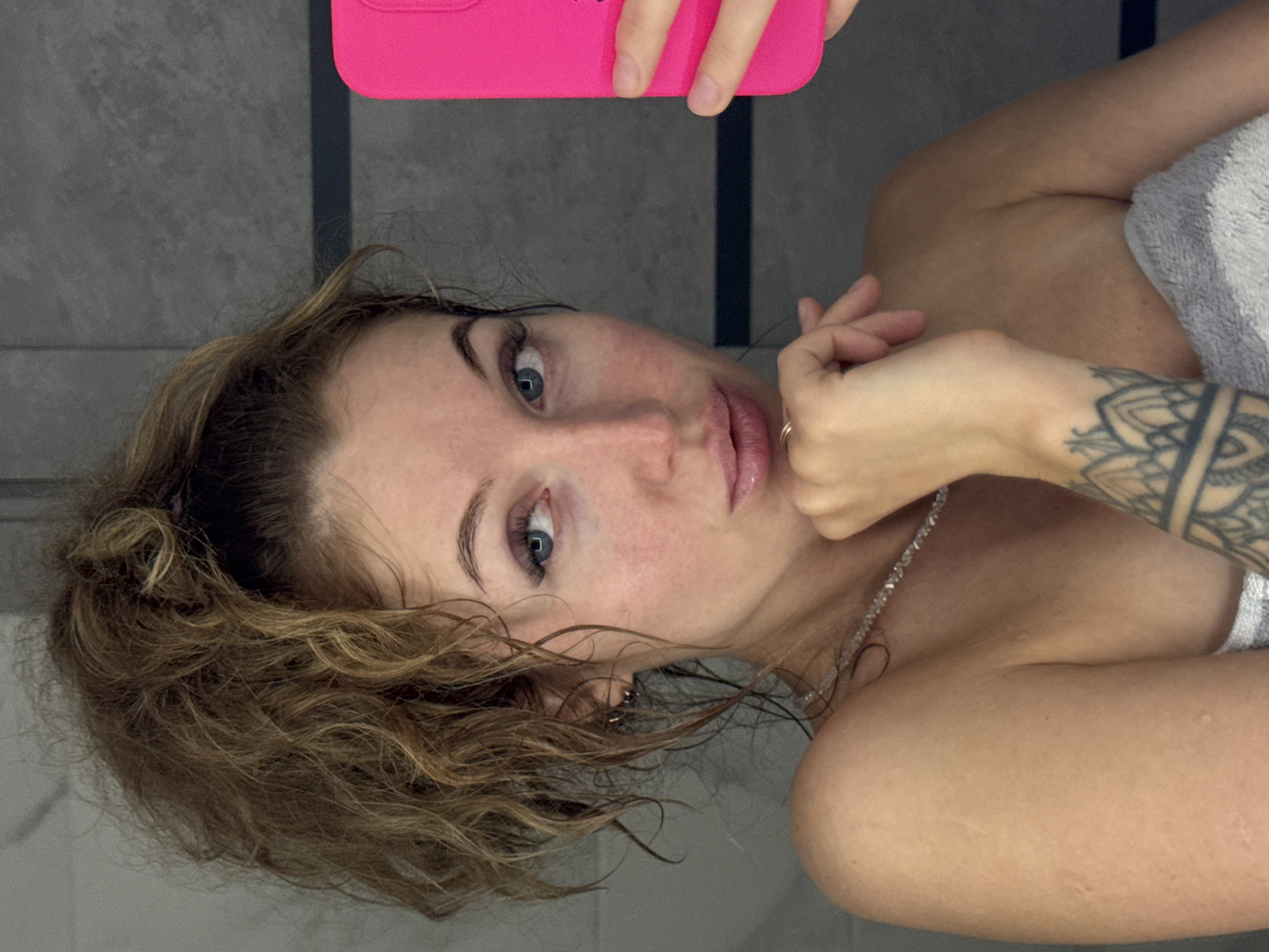 A woman takes a selfie in what appears to be a bathroom setting. She is partially draped in a gray and white striped towel. She holds a pink phone case in her left hand, partially obscuring her face. She has curly, brown hair that is wet near the front. Her eyes are blue and she has a necklace. A tattoo is visible on her right forearm. The background consists of vertically patterned gray wall tiles. There is no evidence of the location being in Chaikovsky, Russia.