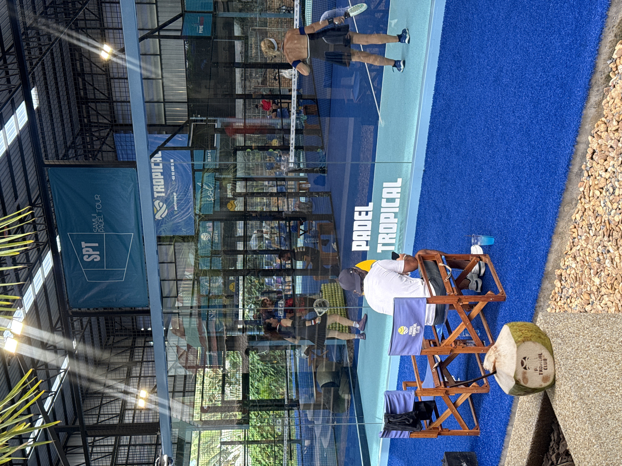 The image depicts an indoor padel court located at Padel Tropical Club in Ban Bang Rak, Thailand. The court has a vibrant blue artificial turf surface and is enclosed by glass walls. Several individuals are visible within and around the court. On the court, two women are actively playing padel, with one in mid-swing and the other positioned defensively. Spectators are also present; a man wearing a white t-shirt and a cap sits in a director's chair in the foreground, looking at his phone. Next to him, another director's chair is occupied by a person whose back is to the camera, and a third chair is empty. A freshly opened coconut with a straw and a logo of "Padel Tropical Club" is placed on a textured surface in the foreground. Overhead lights illuminate the court, casting beams of light. Banners displaying "SPT SAMUI PADEL TOUR" and "PADEL TROPICAL" are prominently displayed. The overall scene suggests a recreational or competitive padel event taking place in a tropical setting.