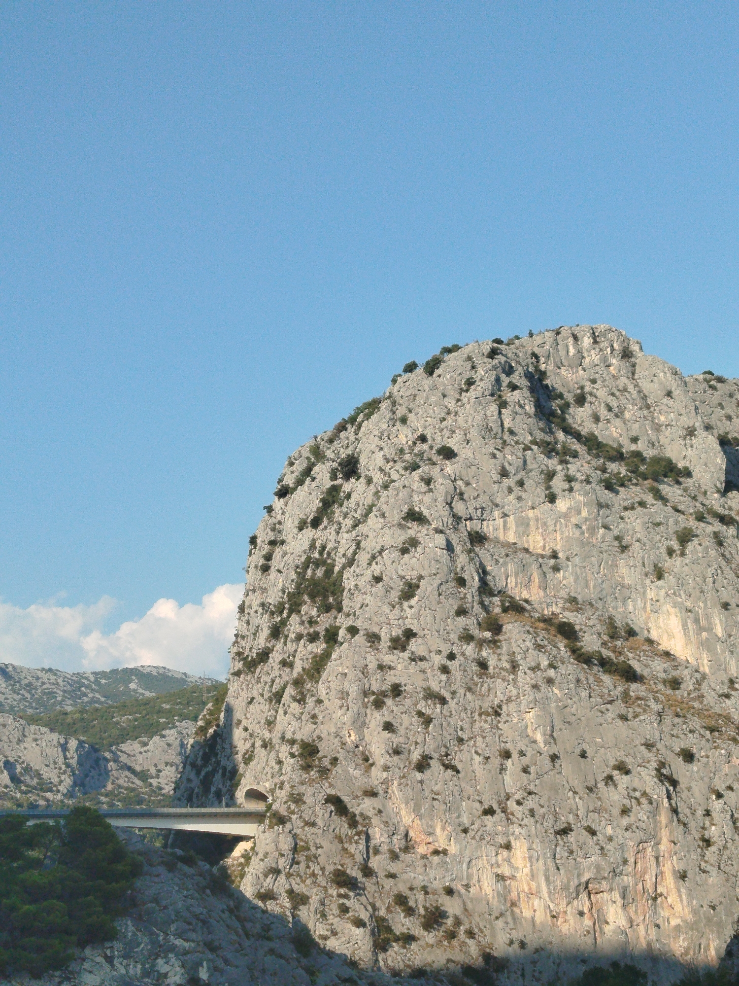 The image captures a vast, rugged, grey rock face dominating the frame, with sparse green vegetation clinging to its surface. A bright blue sky with a few wispy white clouds forms the upper portion of the image. In the lower left quadrant, a modern bridge spans a gap, with a dark tunnel entrance visible within the rock face to the right of the bridge. The landscape suggests a mountainous or canyonous region. This scene is characteristic of the area around the city of Omiš, Croatia, known for its dramatic karst formations and the Cetina River canyon.