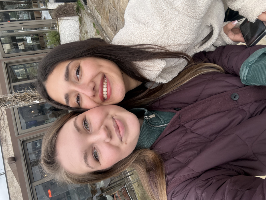 A close-up outdoor photograph depicts two young adult females. The individual on the left has light brown hair and green eyes, wearing a dark plum quilted jacket over a dark green ribbed mock-neck top, and displays a slight smile. The individual on the right has long dark brown hair and dark eyes, wearing a white fuzzy jacket, and exhibits a wide, open-mouthed smile with visible teeth. Her right hand, featuring light-colored manicured nails, holds a black smartphone. Both are looking directly at the camera, with their heads close together. The background reveals the exterior of a commercial building, likely a restaurant or cafe, characterized by large glass windows and doors, a brown upper facade, and leafless vine structures. To the right, a white textured wall with green foliage and a paved ground surface are visible. The sky is overcast. While the general urban setting aligns with a city like Budva, Montenegro, no specific landmarks or identifying features are present in the image to pinpoint an exact location within Budva.