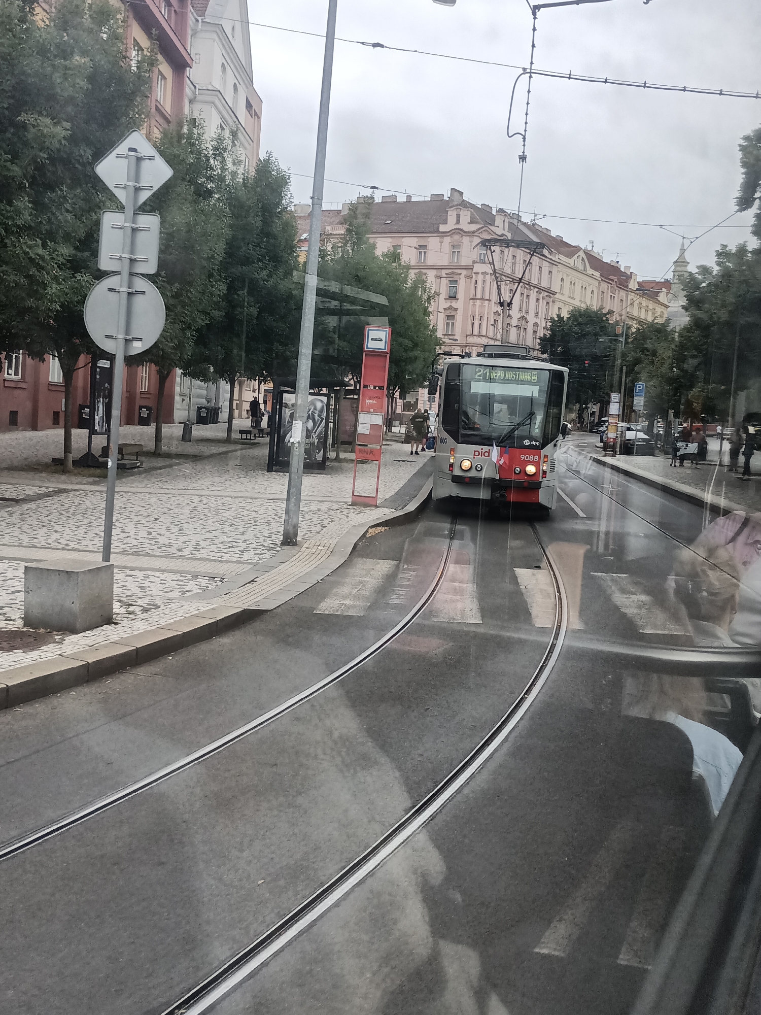 This media file captures a street scene in Prague, Czechia, on a cloudy day. The primary focus is a modern tram on tram tracks, moving down a wet street. The tram is white with red accents and displays the number "21" and the destination "JEPO HOSTIUNR" on its front. It also has the "pid" logo and the number "9088" visible. The street is paved with cobblestones in some areas, and there are trolley wires overhead, typical of European cityscapes.
In the background, buildings with characteristic European architecture line the street. Trees with full foliage add to the urban greenery. Several people are visible, including individuals waiting at a tram stop, a person walking on the cobblestone path, and figures reflected in the tram's windows. The wetness of the road suggests recent rain or ongoing damp conditions.
To the left of the tram tracks, there is a bus stop shelter with advertising posters. A set of traffic signs, including a diamond-shaped sign, are also present. The overall mood of the image is somewhat subdued, likely due to the overcast weather. The scene depicts a typical urban commute in Prague, with the tram as the central mode of transport.