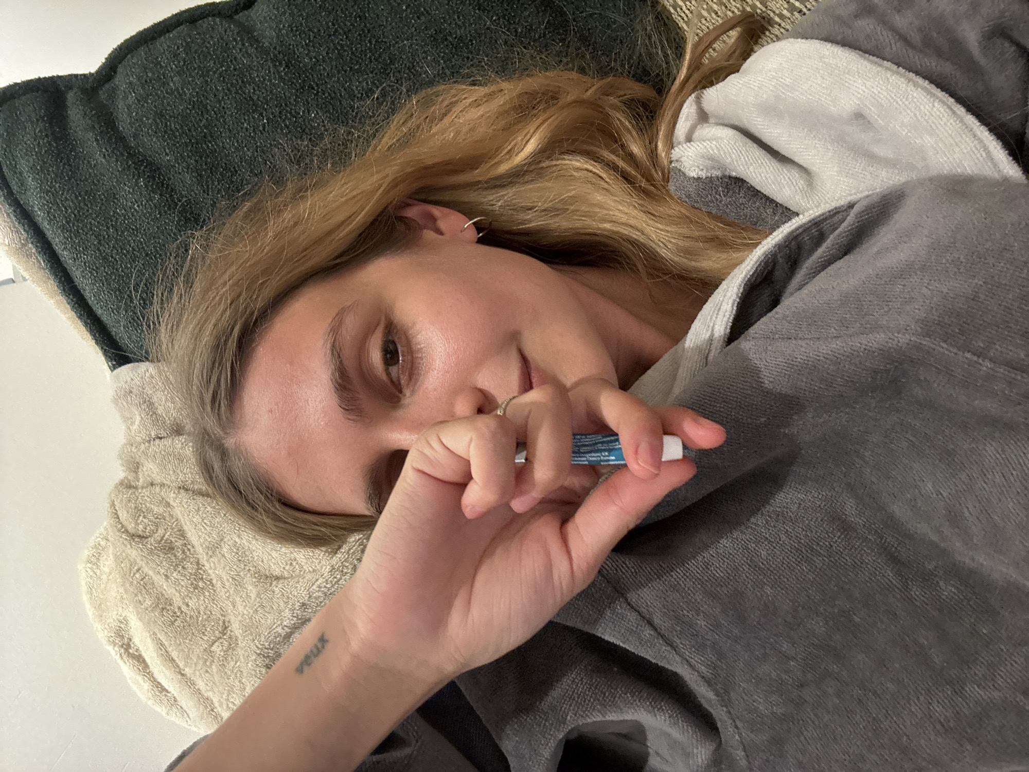 A person lies on cushions, covered by a grey robe with a white hood. The person holds a small, blue and white tube in their right hand. The person's hair is blonde and pulled back from their face. The location is unknown.