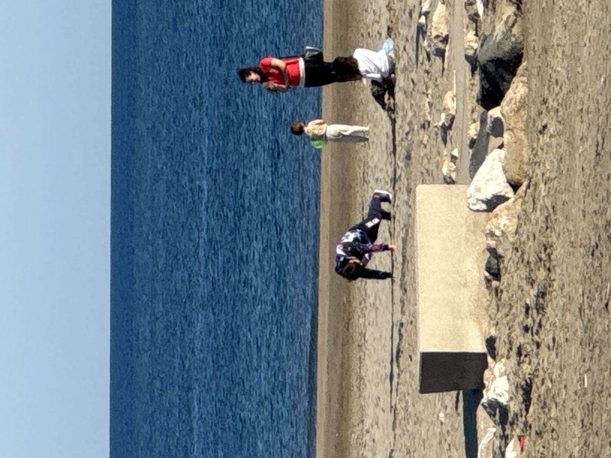 The image, taken in Nettuno, Italy, captures a seaside scene with several individuals engaged in different activities on a bright, clear day. The vibrant blue sea occupies the left side of the frame, appearing calm with subtle ripples on its surface.
Along the right edge of the sea, a light brown sandy or rocky shore extends. Further inland, the terrain becomes more rugged with numerous light-colored boulders and rocks scattered across the sand. A long, rectangular concrete block or barrier is visible in the mid-ground on the right side of the shore. The sky, a pale blue, is visible above the sea, indicating fair weather.
Three individuals are present on the shore. Closest to the water, a person wearing a red top and dark pants stands, seemingly observing the sea. A white cloth or item is visible on the sand near this person. Behind them and slightly further from the water, a smaller individual, likely a child, stands wearing a light-colored and possibly patterned top with light pants, and appears to have a green object around their waist or held in their hand. Further to the right, near the concrete block, another person in a dark, multi-patterned top and dark pants is seen in a bent-over or push-up like position, suggesting they are exercising or stretching.
The overall impression is one of leisure and activity by the coast, likely during daytime given the clear conditions and bright sunlight. No visible text is present in the image.