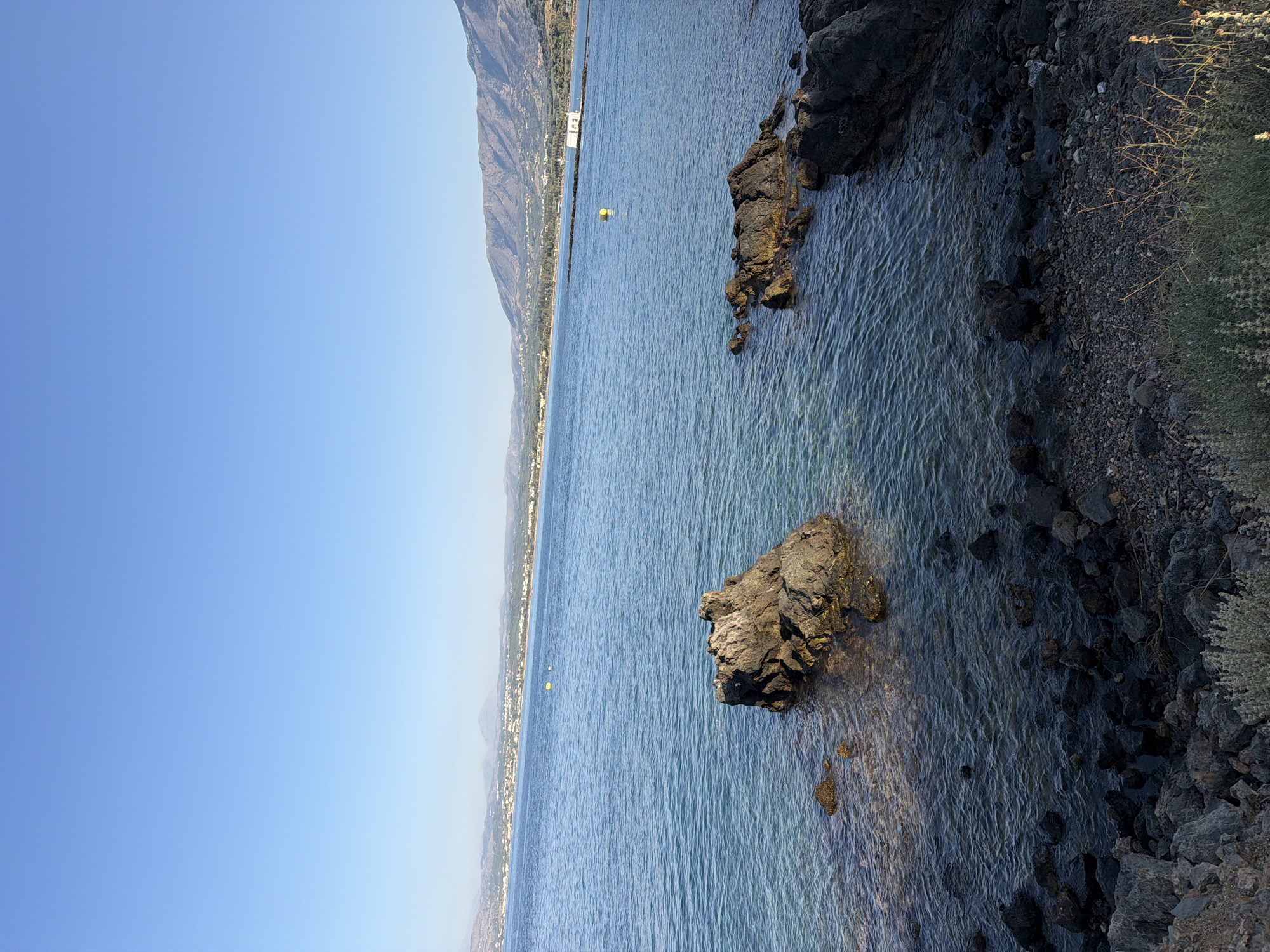 The image shows a coastal scene in Georgioupoli, Greece. In the foreground, a rocky shore transitions into clear blue water. Several dark rocks emerge from the sea, with one prominent, jagged rock in the mid-left of the frame. The water is calm, with gentle ripples reflecting the clear blue sky above. In the distance, a coastline stretches across the horizon, featuring a low-lying town or village with buildings nestled amongst green vegetation. Mountains rise behind the settlement, their slopes covered in sparse greenery. Two yellow buoys are visible in the water, one further out than the other. A white structure, possibly a building or platform, is situated on the far right of the distant coastline. The overall scene is bathed in natural daylight, suggesting it was taken during the daytime. There are no people visible in the image.