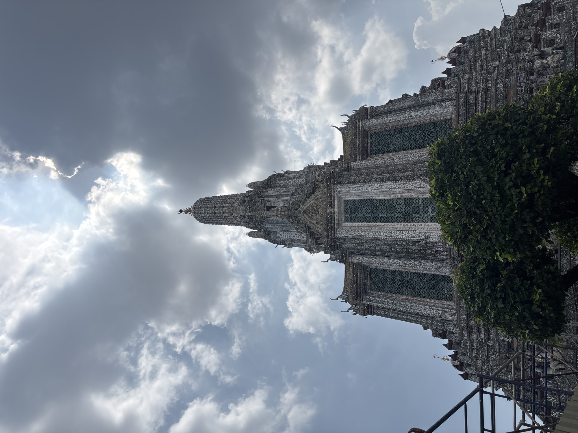 The image shows Wat Arun Ratchawararam Ratchawaramahawihan (Temple of Dawn), a Buddhist temple in Bangkok, Thailand, on the west bank of the Chao Phraya River. The temple's central prang (tower) is prominently featured against a backdrop of a cloudy sky with sunlight breaking through. Part of a tree and a metal railing are visible in the foreground to the right and the bottom right corner respectively. The temple's intricate detailing, including its mosaic tiles and ornate carvings, can be observed.