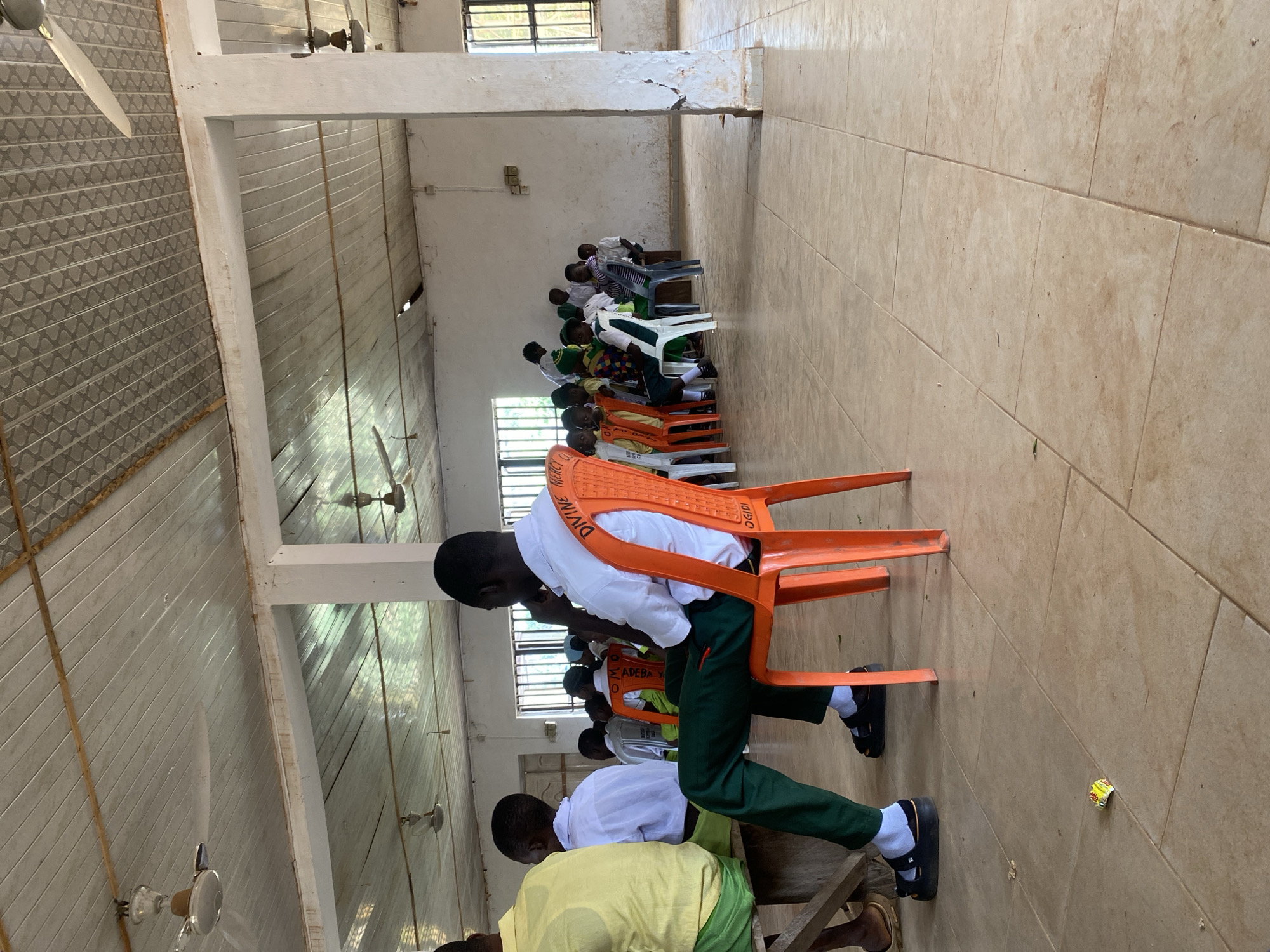 An indoor scene depicts a gathering of people, primarily young individuals dressed in school uniforms, seated in a room with a tiled floor and a suspended ceiling with multiple ceiling fans. The room appears to be a hall or assembly area, with large windows on the right side letting in natural light. In the foreground, a young male wearing a white shirt and green pants sits in an orange plastic chair, facing away from the camera. Other individuals are visible in the background, some seated on benches and others on chairs of various colors, including white and green. A few adults are also present, some standing. The environment is simply furnished, with an emphasis on seating. The overall impression is of a communal gathering or event, likely school-related, taking place in a functional, open space.