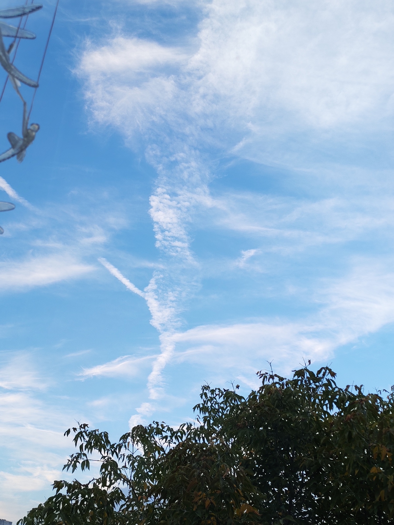 An outdoor, low-angle shot captures a bright blue sky with wispy white clouds. In the upper left corner, a part of a sculpture is visible, featuring metallic, abstract shapes that resemble elongated figures or wings, with one element appearing to be a stylized human form suspended from above by a thin wire. Below the sculpture, the branches and dark green leaves of a large tree fill the lower portion of the frame, obscuring the ground and any potential urban landscape. A faint, thin white line, possibly a contrail, is visible in the middle distance of the sky. The image does not contain any identifiable landmarks that would indicate its specific location within Izhevsk, Russia.