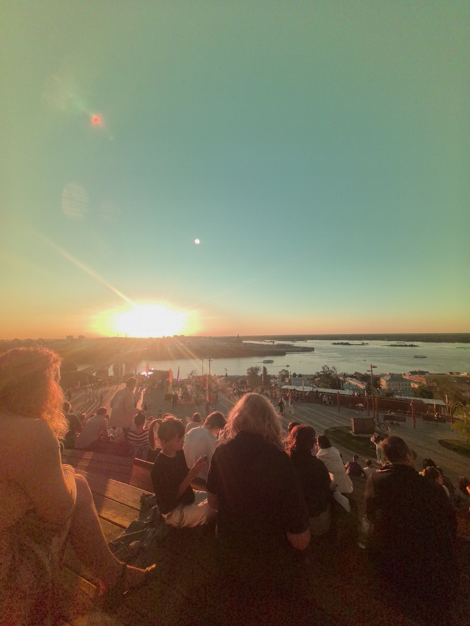 The image shows a crowd of people gathered on a hill overlooking a wide river and a city skyline at sunset. The sun is low on the horizon, casting a warm glow over the scene. In the foreground, people are seated on what appears to be a tiered wooden structure, facing the river and the city. Some individuals are looking towards the sunset, while others are interacting with each other. The river is dotted with boats, and buildings line the opposite bank. The sky is a gradient of orange and blue, with the sun's rays prominently visible. This appears to be a public gathering or event, possibly a concert or festival, taking place in Nizhny Novgorod, Russia, given the characteristic layout of the Volga River and the city's architecture. The overall atmosphere is one of relaxation and communal enjoyment of the sunset.