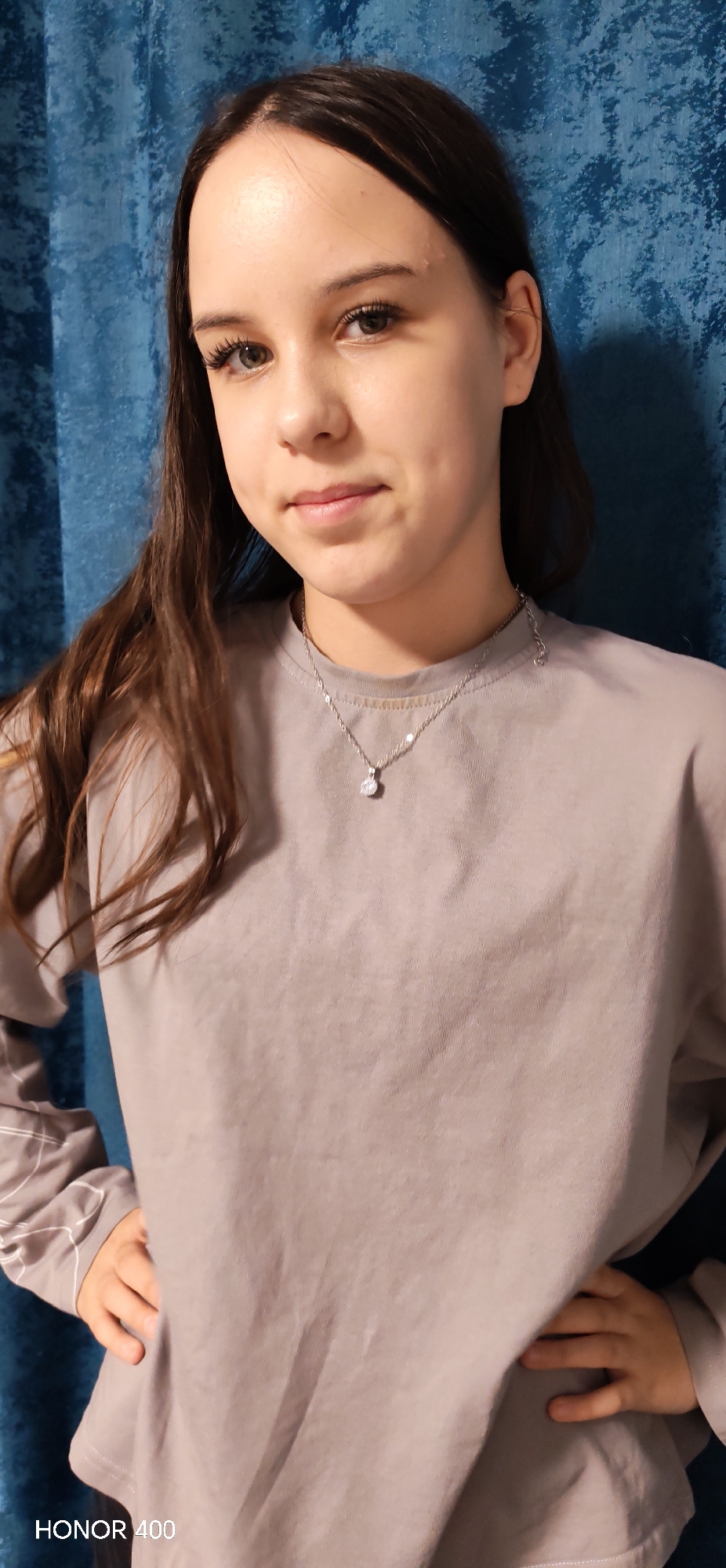 A young woman stands indoors against a blue curtain. She has brown hair and wears a light gray long-sleeved shirt and a silver necklace with a single diamond pendant. Her hands are on her hips. The location is Noviy, Russia. The make of the camera used to take the picture is "HONOR 400".
