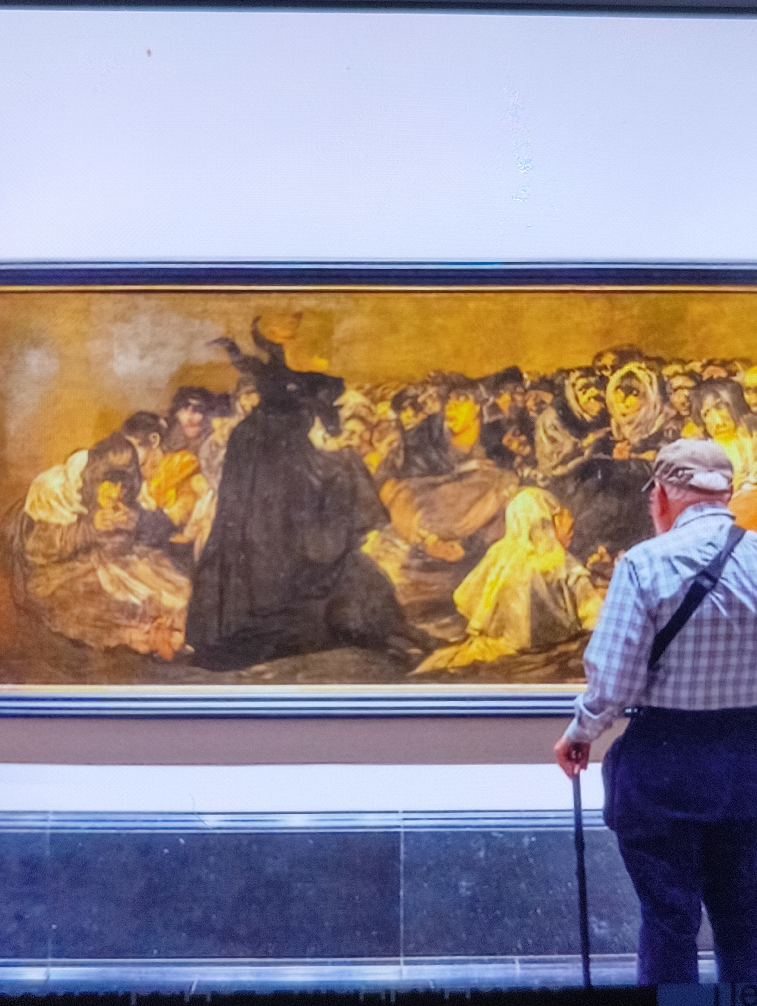 An elderly man with a cane is observing a painting in a museum. The painting depicts a scene with a central figure in a black cloak with horns, surrounded by a crowd of people in various states of distress or awe. The man is wearing a plaid shirt and dark trousers, and a dark strap is visible across his chest. The painting itself is framed and hung on a white wall, with a dark base below it. The lighting in the room is subdued. The image is taken from a slightly elevated angle, looking down at the man and the painting.