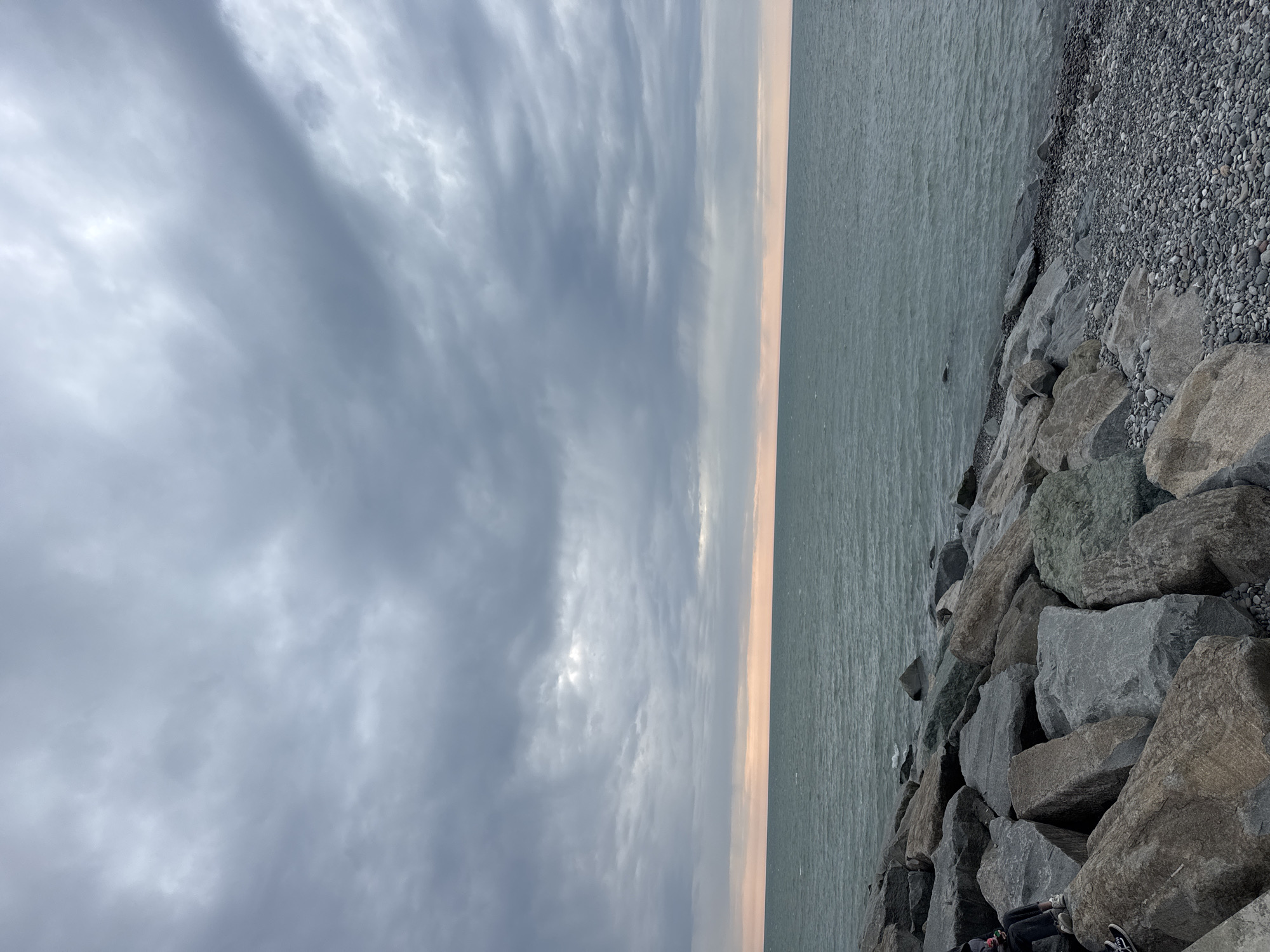 The image depicts a landscape with a body of water on the right, a rocky shoreline in the lower right corner, and a cloudy sky dominating the left side. The horizon line features a thin strip of muted orange or pink light, suggesting sunrise or sunset. The rocks are various shades of gray, with some pebbles and smaller stones mixed in. The water appears calm and slightly reflective. The clouds are layered and varied in density, with some areas showing patches of lighter sky. The location is Unknown, Unknown.
