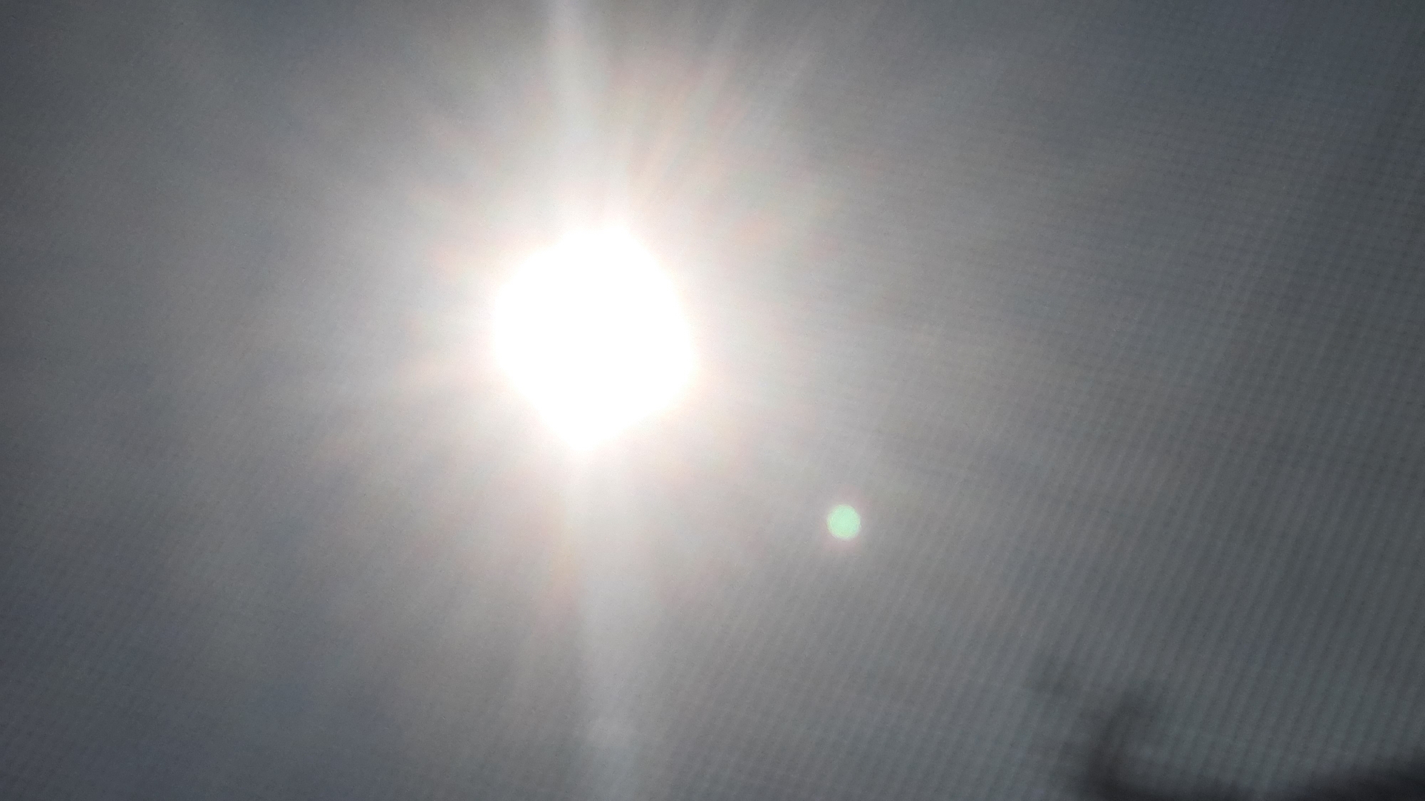 The image shows a bright sun with visible rays against a hazy, grey sky. There is a faint, circular green orb to the left of the sun, likely a lens flare or reflection. The bottom left of the image shows a dark, indistinct silhouette which could be a tree branch or other object obscuring the view. The image appears to have been taken indoors, possibly looking out a window or through a screen, as indicated by a visible grid pattern across the entire frame. The location is in Shklov, Belarus.