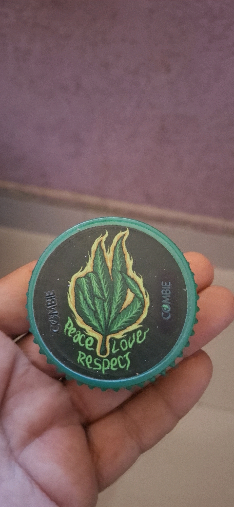 A close-up, vertical shot shows a person's hand holding a green bottle cap with a black sticker on it. The sticker features a drawing of a cannabis leaf with orange flames around it. Below the leaf, the words "Peace Love Respect" are written in green, stylized lettering. The word "COMBIE" is printed vertically in white letters on the left and right sides of the sticker, with a small circular logo between the letters on the right side. The background of the image consists of a blurred purple wall with a thin horizontal line above it. The location is not specified in the image.