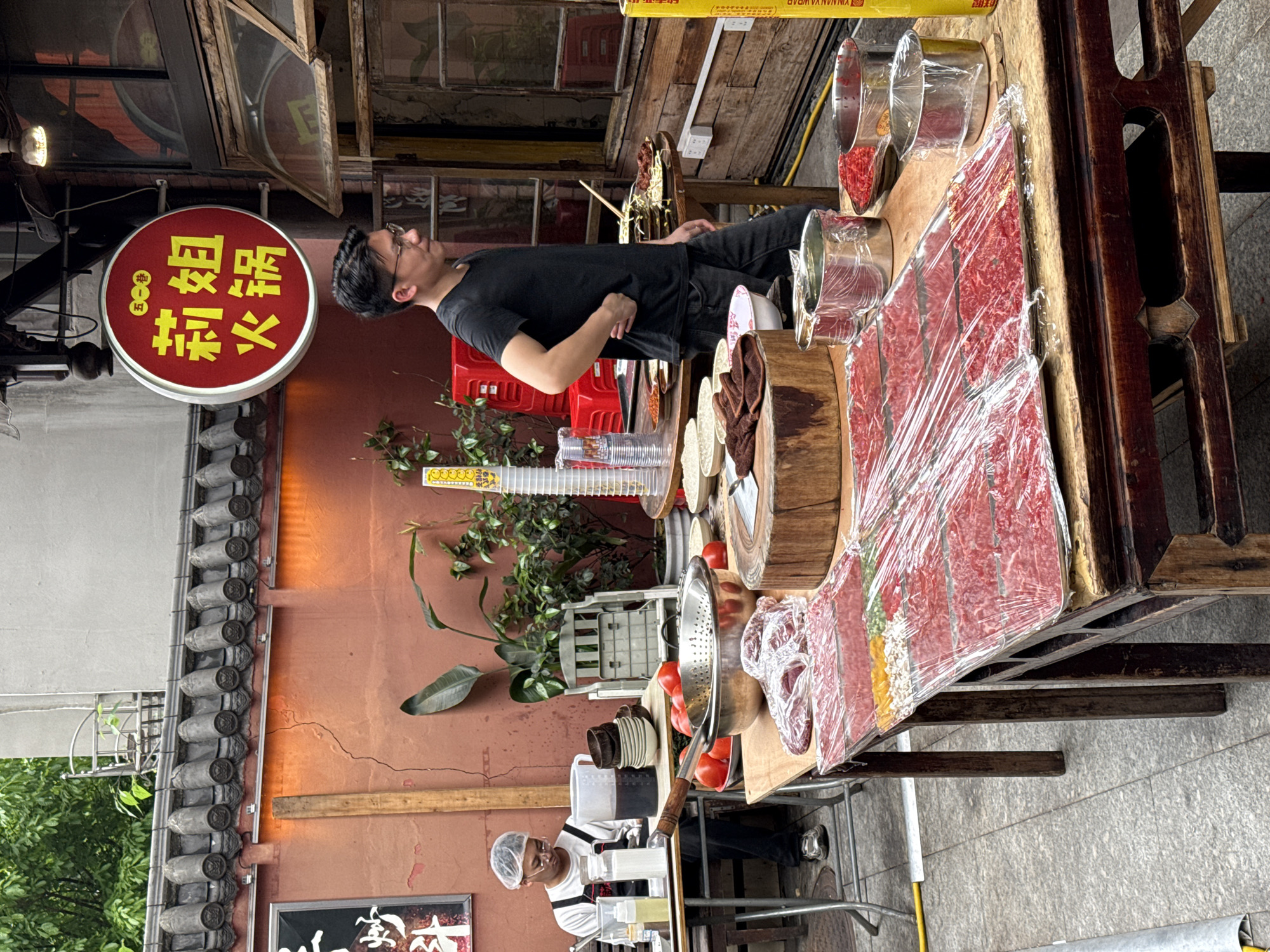 A street scene in Yuzhong District, China, shows a food stall with prepared food. A man in a black T-shirt and jeans is standing to the left of the stall, looking up at a red, round sign with Chinese characters. Another person, wearing a white apron and hair net, is operating another food stand in the lower-left. On the food stall, food is displayed on a large wooden table, including what appears to be meat covered in plastic wrap, along with containers of spices. The stall has red tomatoes, and cut wooden chopping boards, indicating the preparation of the food, possibly street food being prepared and sold in the area. The building behind the stall has exposed roof tiles.