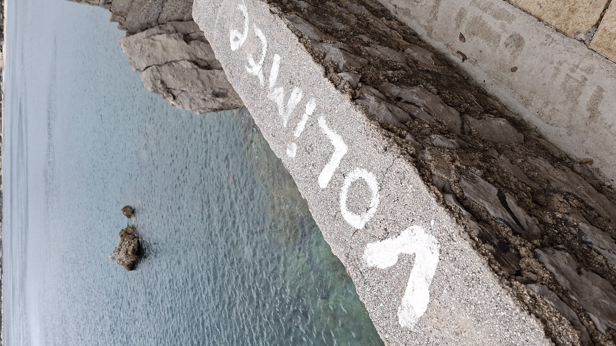 A ground-level shot shows a stone wall with the word "Volice" painted vertically in white on its top edge, overlooking a body of turquoise water. To the left, rocky formations jut out into the water. A small, rocky islet is visible in the water. The wall appears to be part of a structure bordering the water's edge, likely a bridge or pier. The scene suggests a coastal setting. The location is Budva, Montenegro.