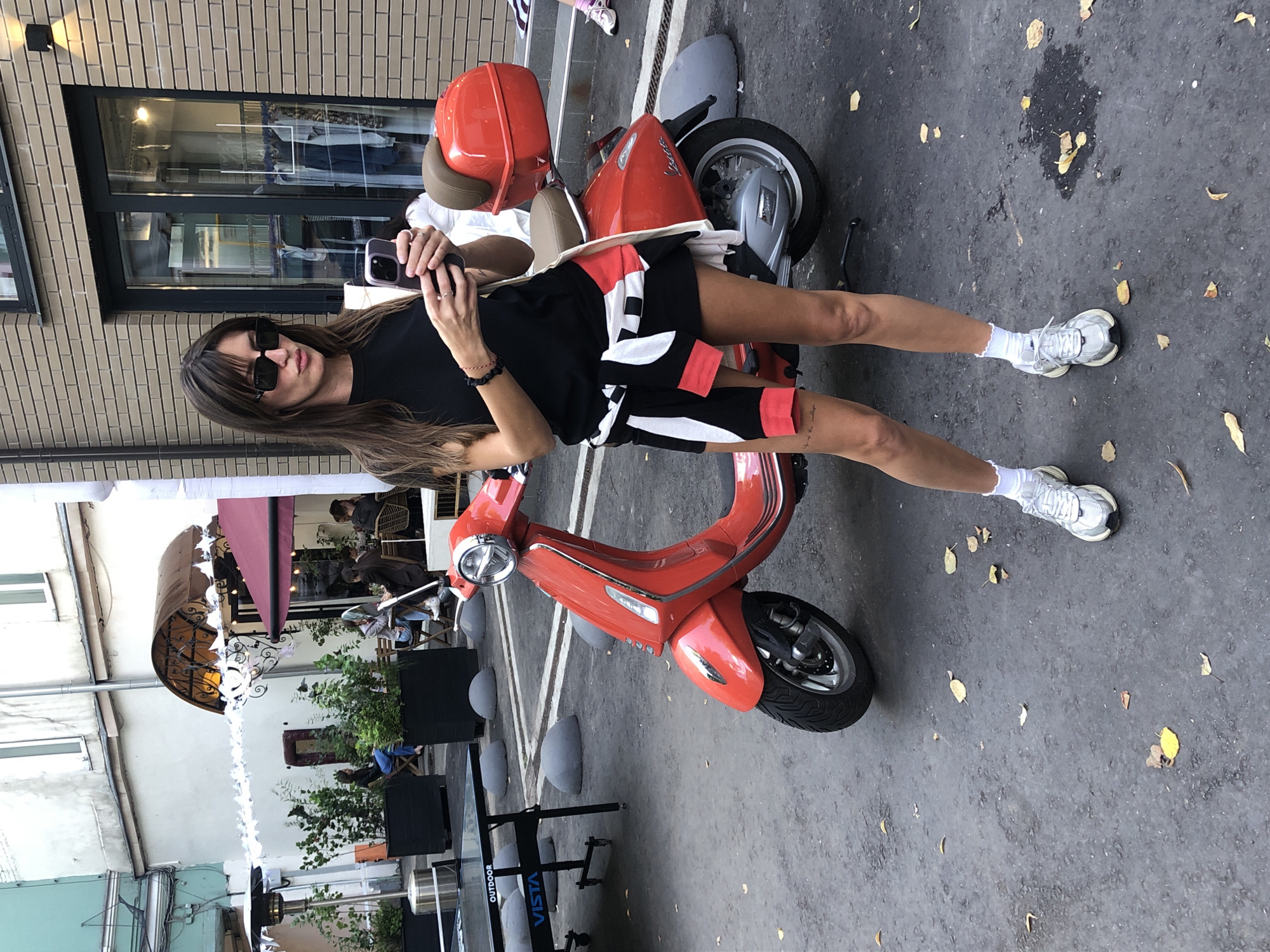 A woman stands next to a red scooter in an outdoor urban setting, possibly a courtyard or alleyway. She is wearing a black dress, white socks, and silver sneakers, with a patterned sweater tied around her waist. She is holding a smartphone and looking at it. In the background, there is a building with a brick facade, a glass storefront, and outdoor seating with umbrellas and plants. A ping pong table is visible to the left. The ground is paved with asphalt and scattered with fallen leaves. The overall scene suggests a casual, possibly commercial or recreational urban environment.