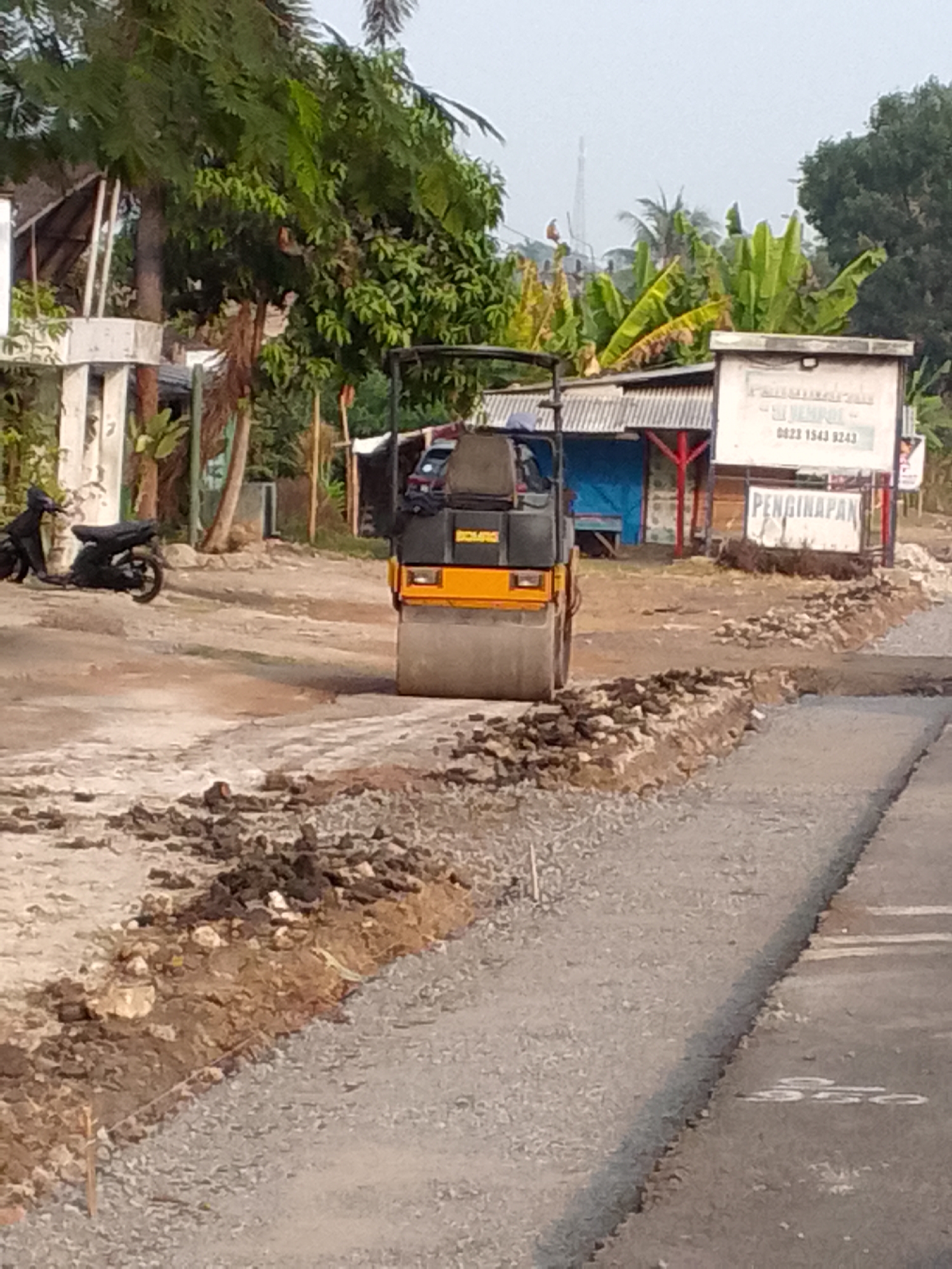 In the city of Mandalasari, Indonesia, an outdoor construction scene is captured. A yellow and black vibratory roller is compacting gravel on a road improvement project. The roller is positioned on the right side of the frame, actively working. To the left, a black motorcycle is parked near a white pillar and some lush green trees. In the background, a small building with a corrugated roof and blue siding is visible, along with a sign that appears to indicate lodging ("PENGINAPAN"). The environment suggests a sunny day with a slightly hazy sky. The road itself shows signs of ongoing work, with excavated earth and loose stones bordering the newly compacted area.