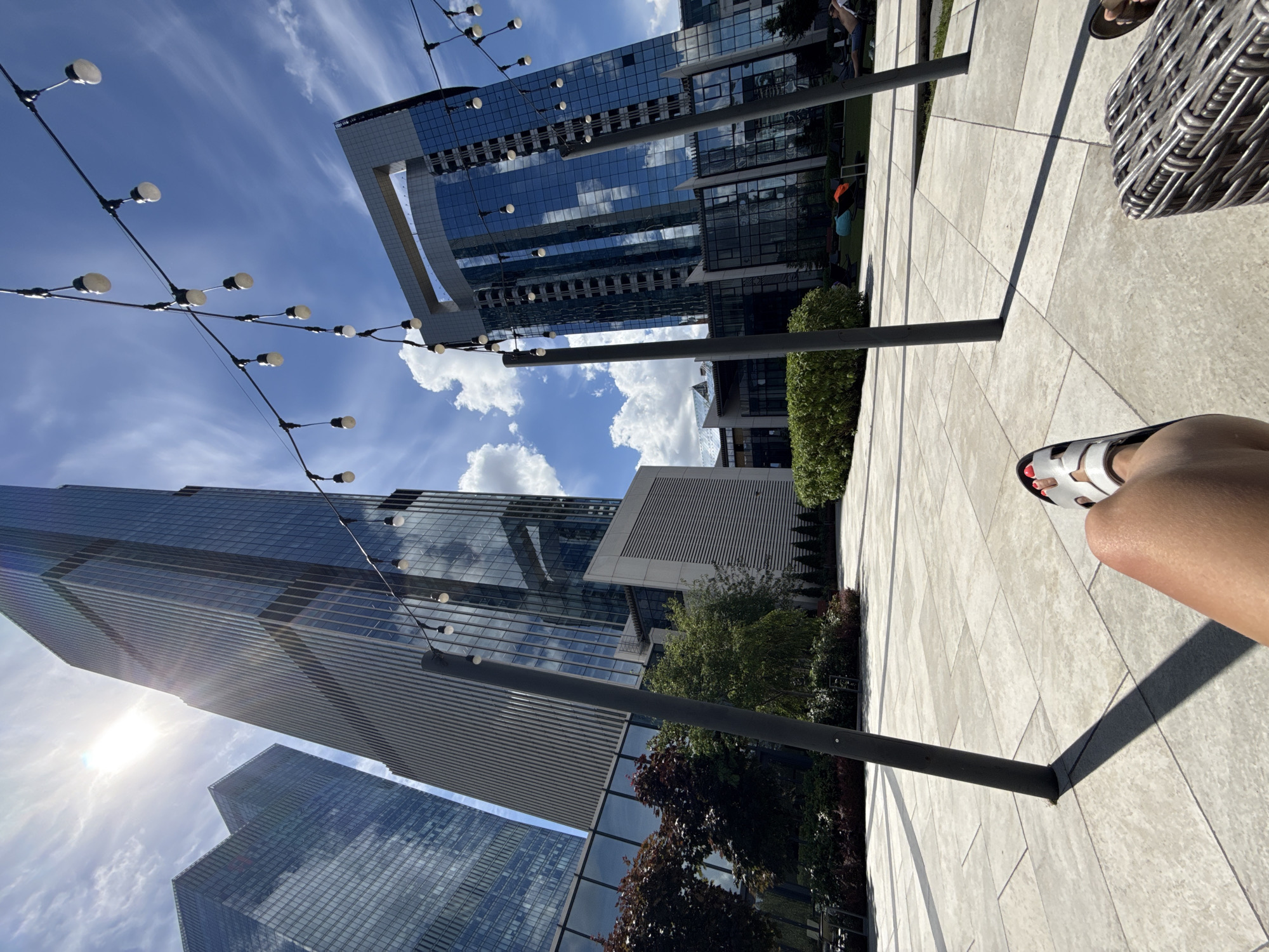 The image shows a sunny day with a blue sky filled with white clouds, in front of modern glass skyscrapers in Moscow, Russia. The perspective is from a person sitting on a chair, with their legs and feet visible in the foreground, wearing white sandals. String lights with exposed bulbs are hanging above, and dark poles are scattered across the tiled ground. In the background, lush green bushes and trees are planted, and several people are seen relaxing on lounge chairs on a grassy area. The architecture of the buildings is sleek and contemporary, with reflective glass facades.