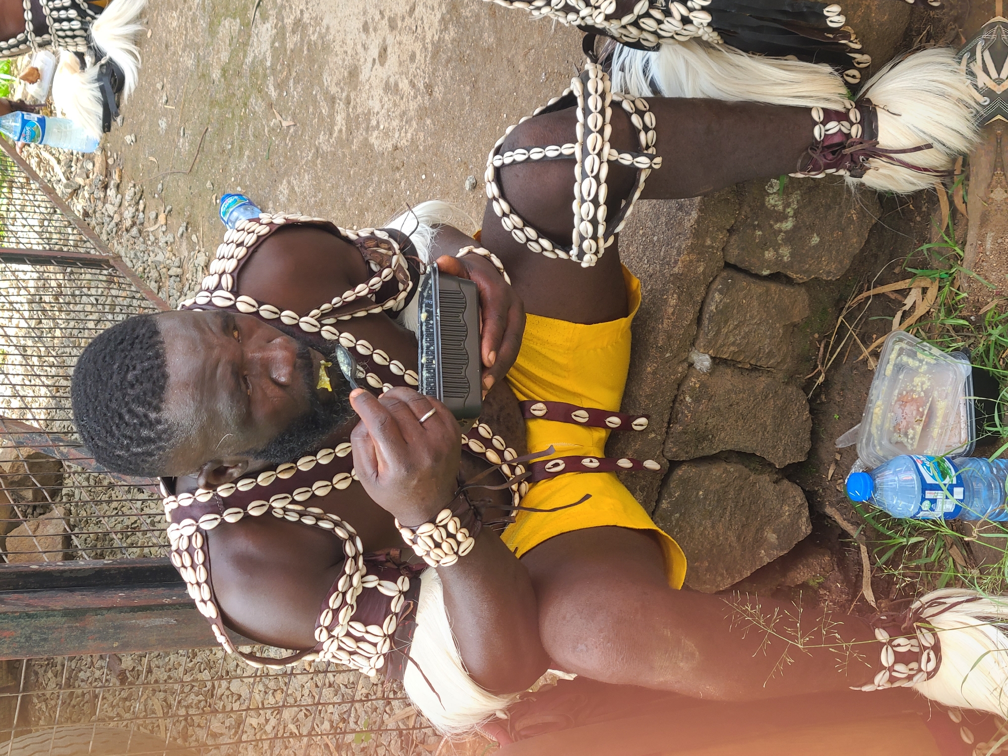 The media file depicts a person adorned in traditional attire, seated on the ground. The individual is wearing a headdress and body ornaments made of shells and what appears to be white horsehair or similar material. Their torso is covered by a garment with shell embellishments, and they are wearing a yellow wrap or skirt. The person is holding a black electronic device and has a ring on their left hand.
The scene is outdoors, with a rough, gravelly ground surface and some rocks. There is a metal fence in the background. Two plastic bottles of water are visible, one in the foreground and another further back. A clear plastic container with some food residue is also present. The lighting suggests it is daytime.
The person's expression is focused, possibly engaged in using the electronic device or observing something off-camera. The overall impression is of a cultural or ceremonial context, given the elaborate traditional clothing. The location cues, such as the vegetation and the type of ground, are consistent with an outdoor setting in Nigeria, specifically Jos, as indicated. No specific time of day or weather conditions are explicitly evident beyond daylight.