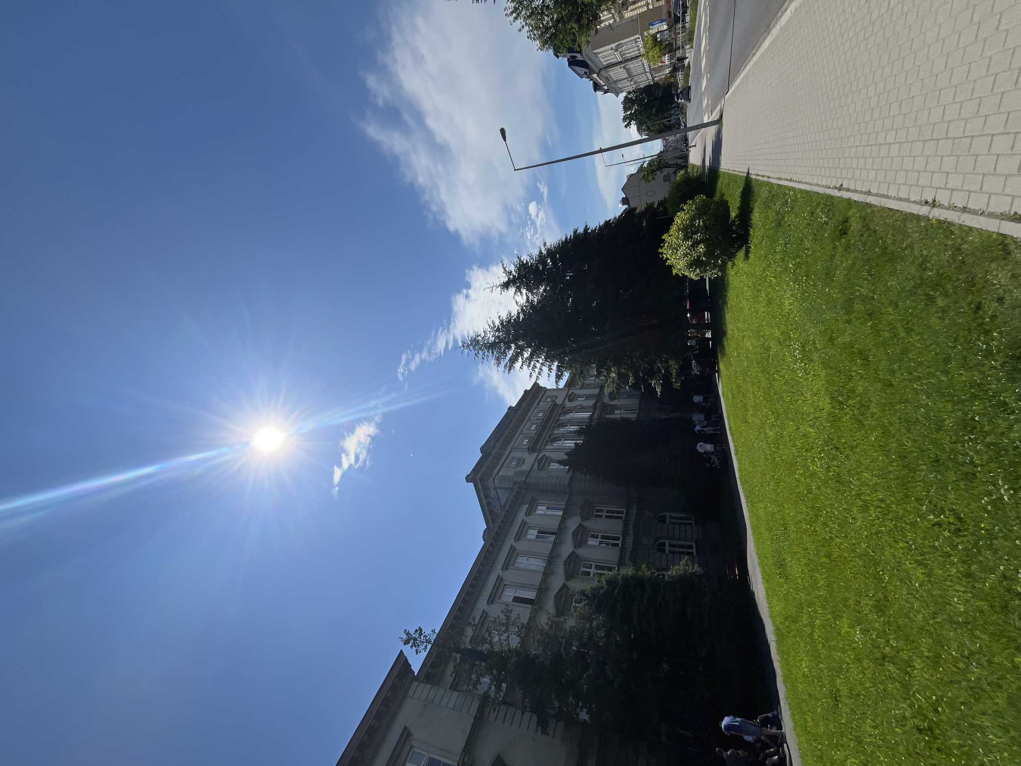 An eye-level, wide-angle shot captures a sun-drenched street scene in an urban environment. The bright sun, positioned in the upper left quadrant of the sky, emits strong rays and lens flare. A large, ornate building with multiple windows and a classical architectural style occupies the left side of the frame. In front of the building, a well-maintained green lawn slopes downwards, with several people visible on the lawn and along a paved walkway. Tall, dark green conifer trees are interspersed with the building and the lawn. To the right, a paved sidewalk runs parallel to a street with a pedestrian crossing. Several cars are present on the street, and more buildings line the opposite side of the road. The sky is a clear blue with scattered white clouds.