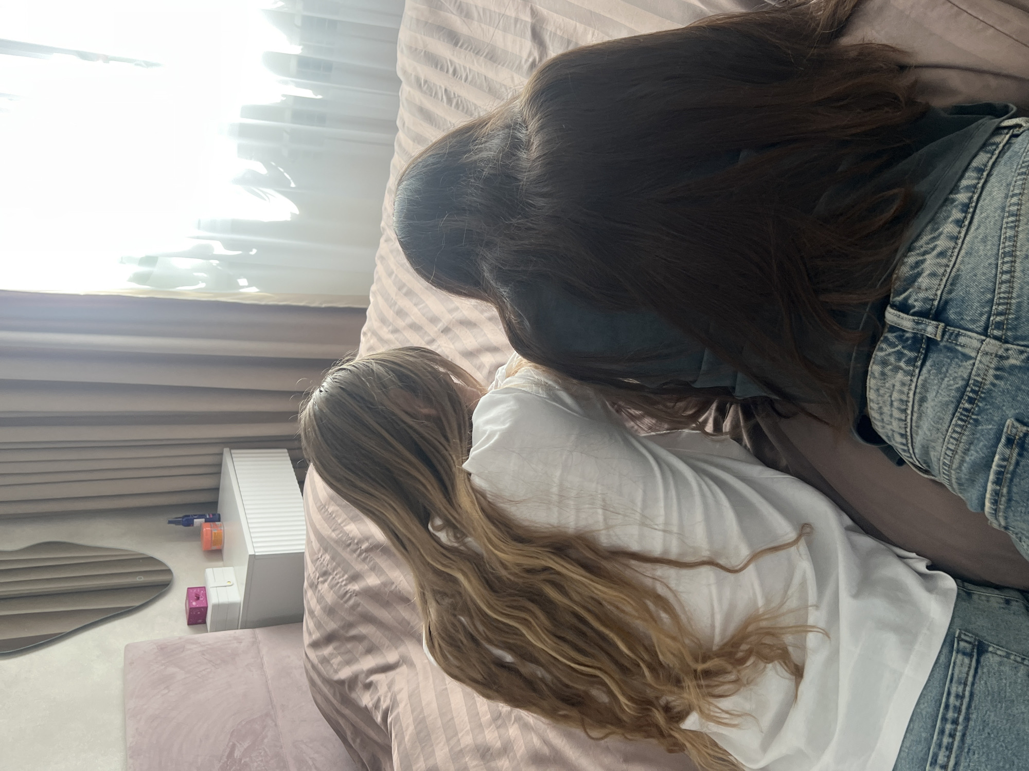 Two individuals, likely women, are shown lying on a bed from a high, diagonal perspective. The person in the foreground has long, wavy, blonde and light brown hair that spreads across the striped, light pink or beige bedding. They are wearing a white top. Behind them, another person is visible, with long, straight, dark brown hair flowing down their back. This individual is wearing a light blue denim jacket or overall over a darker top.
The subjects appear to be resting or relaxing together on the bed. To their left, a brightly lit window with white horizontal blinds allows strong sunlight to stream into the room, suggesting it is daytime. Below the window, there is a light-colored bedside table or shelf holding several small items: an orange container, a pink cube, a white rectangular box, and a small blue object, possibly a pen or applicator. A wavy, organic-shaped mirror is also visible, reflecting the blinds. The overall setting appears to be a cozy bedroom. No visible text is present in the image.