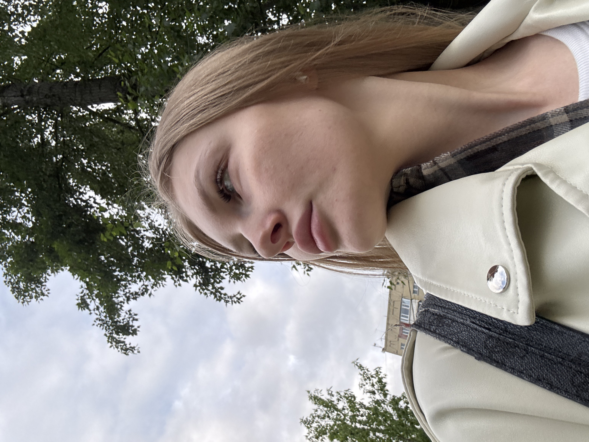 A young woman with blonde hair is depicted from a low-angle perspective, against a background including trees, the sky, and a building. The woman is wearing a light-colored jacket, partially visible, and a plaid shirt. The trees are green, and the sky is overcast. A building with windows is visible in the background. The photo appears to be taken outdoors. Based on the building visible, it is possible that the location is in Perm, Russia.