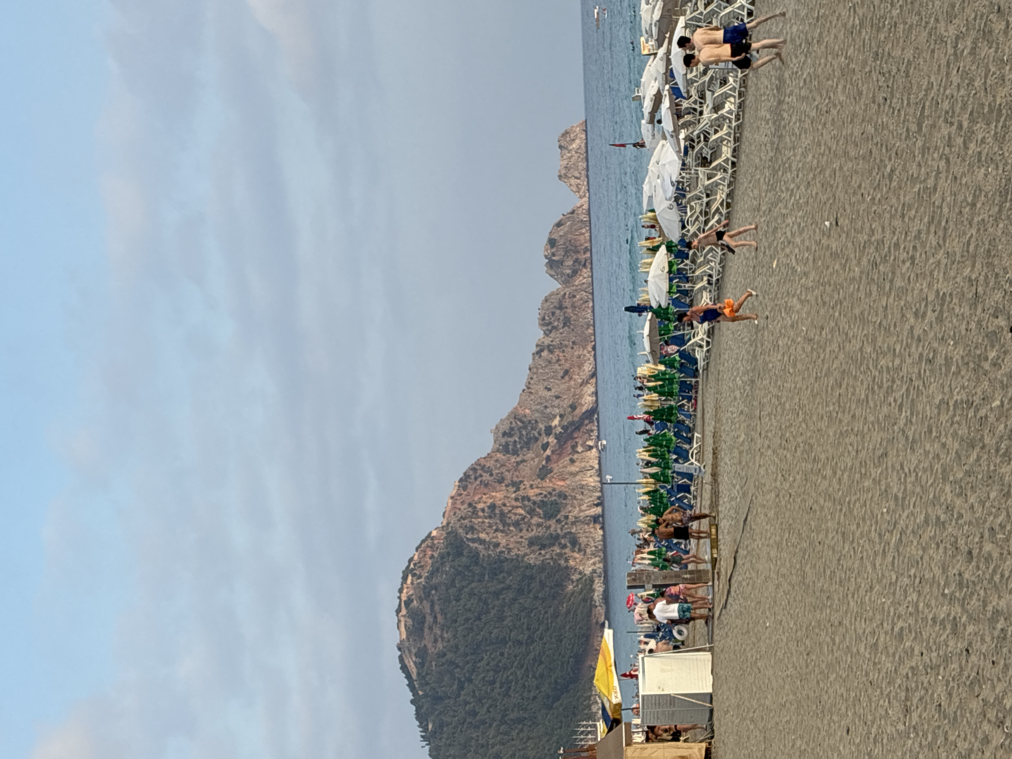 The image depicts a crowded beach in Alanya, Turkey. In the background, a large, verdant mountain with reddish rocky outcrops dominates the landscape, with a fortified structure visible near its peak. The sea is a calm blue, dotted with a few distant boats. The beach itself is covered in sand and lined with numerous rows of lounge chairs and umbrellas, many of which are white and green. Several groups of people are visible on the beach and near the water. In the foreground, two young men in dark shorts walk away from the camera, while to their left, a woman in a blue swimsuit walks with an orange bag. Further left, a group of people, including men in swim trunks and women in patterned shorts, are standing near some beach structures. The sky is a pale blue with scattered clouds. The overall impression is a busy summer day at a popular seaside resort.