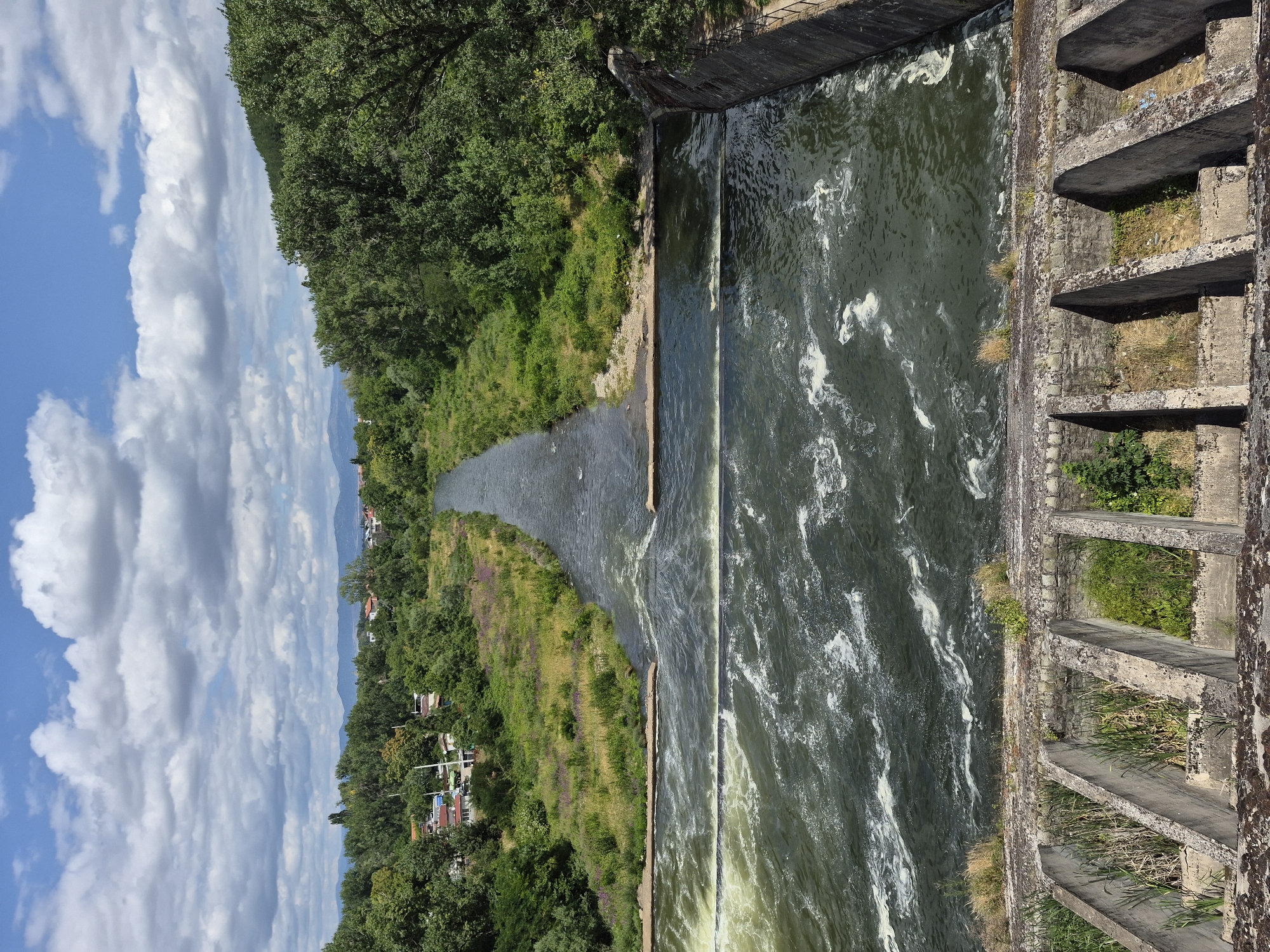 This aerial, eye-level, full shot captures the Pancharevo water dam, a feature of Pancharevo, Bulgaria, on a bright, cloudy day. The dam is constructed of concrete and stone, with visible sections of metal railings along its right edge. The water flowing through the dam exhibits a turbulent, foamy appearance, indicating it is actively moving over the structure. Vegetation, including grass and trees, surrounds the dam and lines the riverbanks, with a small cluster of buildings visible in the distance, nestled amongst the trees on the left bank. The sky is a vibrant blue, adorned with fluffy white clouds, contributing to the overall serene, natural atmosphere of the scene.