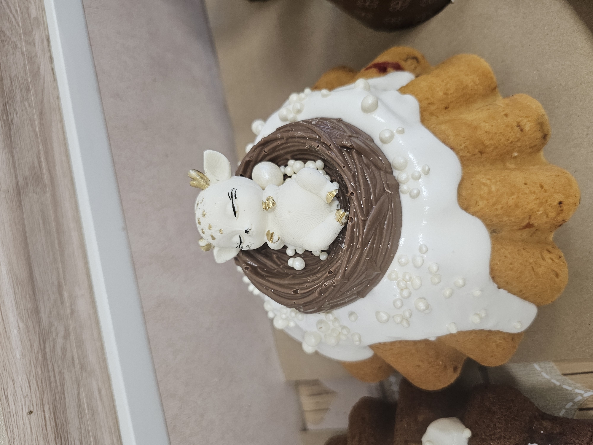 A close-up shot features an ornately decorated dessert. The dessert consists of a cookie-like base, topped with white frosting and small white edible pearls. A chocolate bird's nest, crafted from molded chocolate, is positioned on top of the frosting. Inside the nest, a small, white chocolate deer figurine lies nestled among more edible pearls. The dessert appears to be sitting on a light brown surface near a wooden or faux-wood panel and a white wall or border. There is no evidence to suggest that the dessert is located in Moscow, Russia.