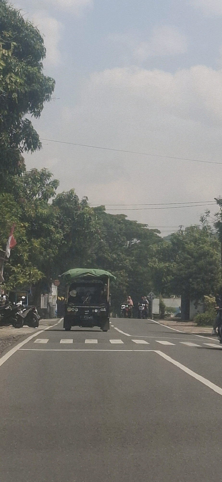 The image captures a street scene in Susukan, Indonesia, under an overcast sky with scattered clouds. In the foreground, a paved road features a pedestrian crossing with white stripes. A dark-colored auto rickshaw, covered with a green tarp, is centrally positioned on the road, facing away from the camera. The rickshaw appears to have a driver and at least one passenger visible.
To the left of the road, parked motorcycles are visible, and a building with signage and a red and white Indonesian flag is situated behind them. Beyond this, dense green trees line the roadside.
Further down the road, several individuals on motorcycles are moving. Two motorcycles are in the middle distance, one with two occupants, and another further back with at least one occupant. To the right, more trees and some white structures, possibly buildings or fences, are present. Power lines are visible stretching across the upper portion of the frame. The overall environment suggests a rural or semi-urban setting.