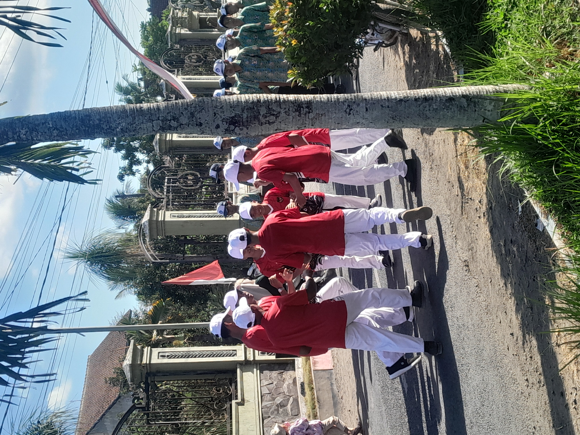 In Sambirejo, Indonesia, a group of young people in matching red shirts and white pants, wearing white hats, are engaged in a procession or performance on a paved street. They are in a circular formation, with some individuals appearing to hold hands and move in a coordinated manner. Behind them, other individuals in patterned shirts and white hats are lined up, suggesting a larger group or parade. A red and white Indonesian flag is visible in the background, near a decorative gate. Palm trees and overhead power lines indicate a tropical, possibly urban or suburban, setting. The environment is outdoors, under a clear blue sky with shadows cast by the sun, suggesting it is daytime.