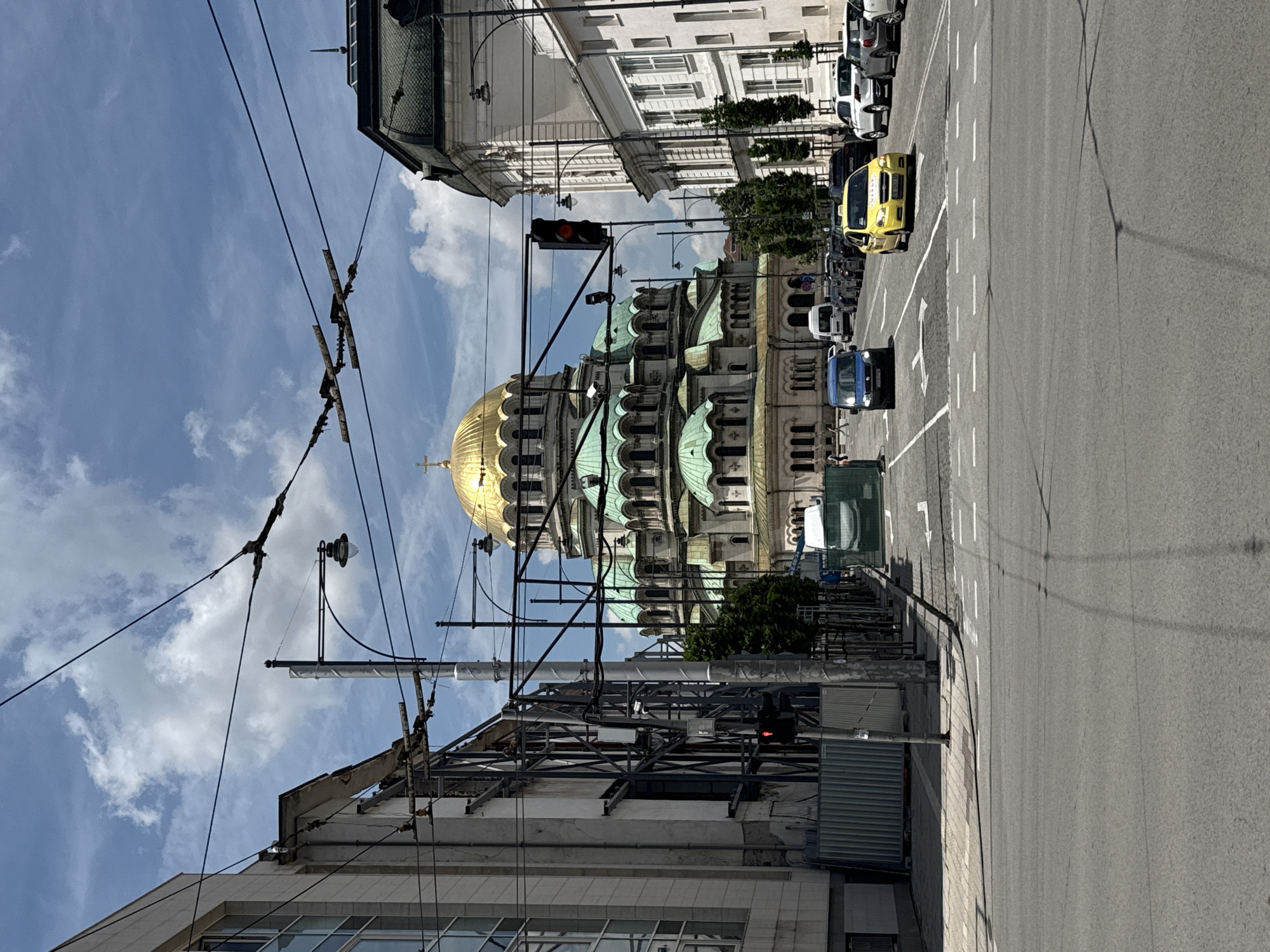 The image displays a daytime street scene in Sofia, Bulgaria, dominated by the majestic Alexander Nevsky Cathedral on the left. The cathedral features a large, prominent golden dome and several smaller green domes, indicative of its Orthodox Christian architecture. Its stone facade exhibits intricate details under the bright sunlight.
A wide asphalt road runs across the foreground and into the distance, marked with white lane lines and directional arrows. Several vehicles are present on the road, including a bright yellow taxi cab on the right and a blue van further down, suggesting an active urban environment. Other cars are parked along the left side of the street.
Above the street, a dense array of black electrical wires and utility poles spans the view, characteristic of a city with trolleybus or tram transport infrastructure. A red traffic light can be seen near the center, indicating current traffic conditions. Alongside the road, multi-story buildings with classic architectural styles are visible, interspersed with some greenery.
The scene is bathed in clear, bright daylight, with a blue sky adorned with scattered white clouds, suggesting good weather. The overall impression is that of a bustling yet calm city atmosphere, highlighting the blend of historical landmarks and modern urban life. No clearly readable text is visible on signs or vehicles, though there appears to be writing on the side of the yellow taxi.