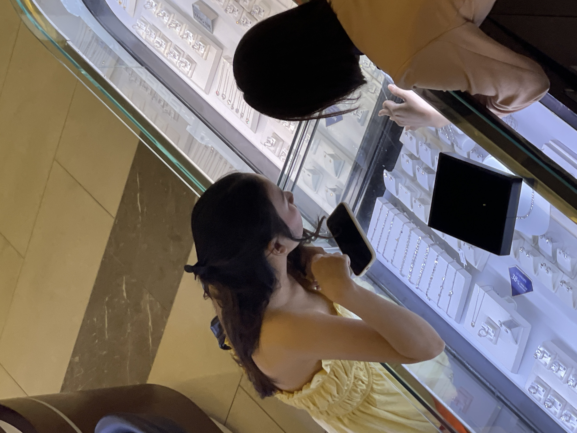 Two people are viewing jewelry inside a shop. One individual, wearing a yellow dress and dark hair, holds a smartphone, apparently taking a photo or video of the jewelry display. The other person, whose back is to the camera, is dressed in light-colored clothing and is also looking at the jewelry. The jewelry is displayed in illuminated cases behind glass. The location is suggested to be in Nha Trang Ward, Vietnam.