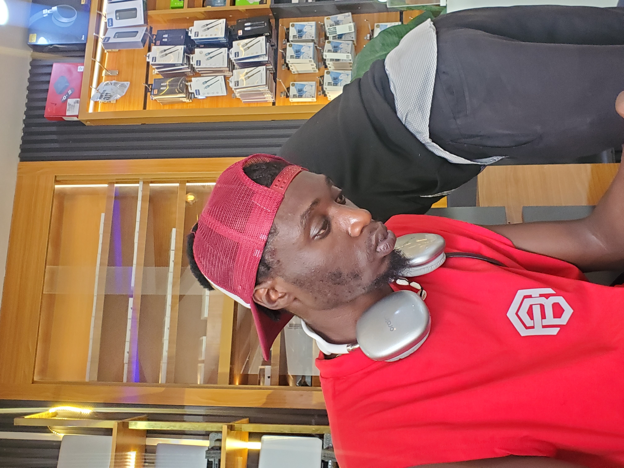 The media file displays a close-up shot of a young Black man from the chest up. He is wearing a red baseball cap and a red t-shirt with a white geometric logo on it. He has silver headphones around his neck. The man is looking to his right with his lips slightly puckered. He appears to be indoors, possibly in a retail setting, with shelves of electronic accessories visible in the background. The shelves are stocked with various boxed items, including what appear to be chargers, cables, and other electronic peripherals. The lighting suggests it is daytime. The overall impression is casual, perhaps a moment captured while the man is browsing or waiting in a store. The location context provided is Jos, Nigeria.
