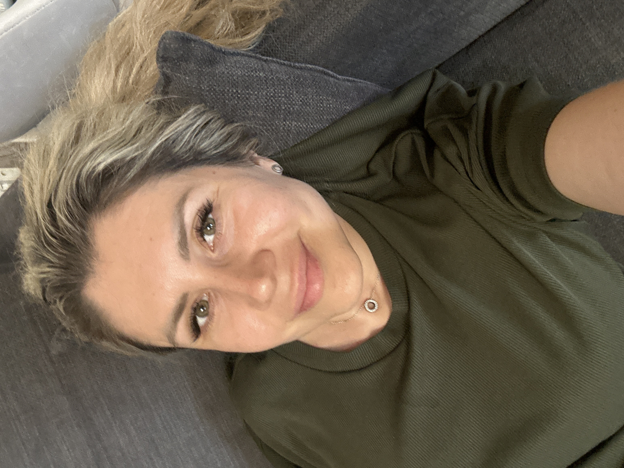 The image shows a close-up, selfie-style shot of a woman lying on a grey couch. She has blonde hair pulled back and is wearing a green, ribbed long-sleeved shirt. Her face is turned towards the camera, and she is smiling. She is wearing light makeup, including mascara and possibly false eyelashes, and a delicate necklace with a small circular pendant. The setting appears to be indoors, likely a living room, given the couch. The lighting is soft, suggesting it could be daytime, but no specific time can be determined. The overall mood is relaxed and casual. No text is visible in the image.
