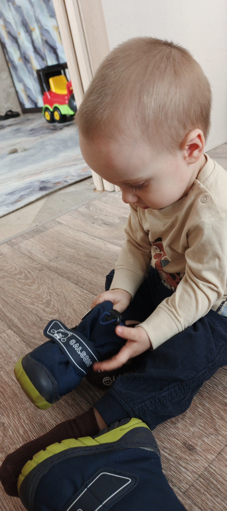 A young child is sitting on a wooden floor in what appears to be an indoor setting, possibly a home. The child is wearing dark blue pants, a light beige long-sleeved shirt, and is in the process of putting on dark blue and yellow shoes with a "Calssic" logo on the strap. In the background, partially visible, is a toy truck on what seems to be an area rug. The location is the city of Chaikovsky, Russia.
