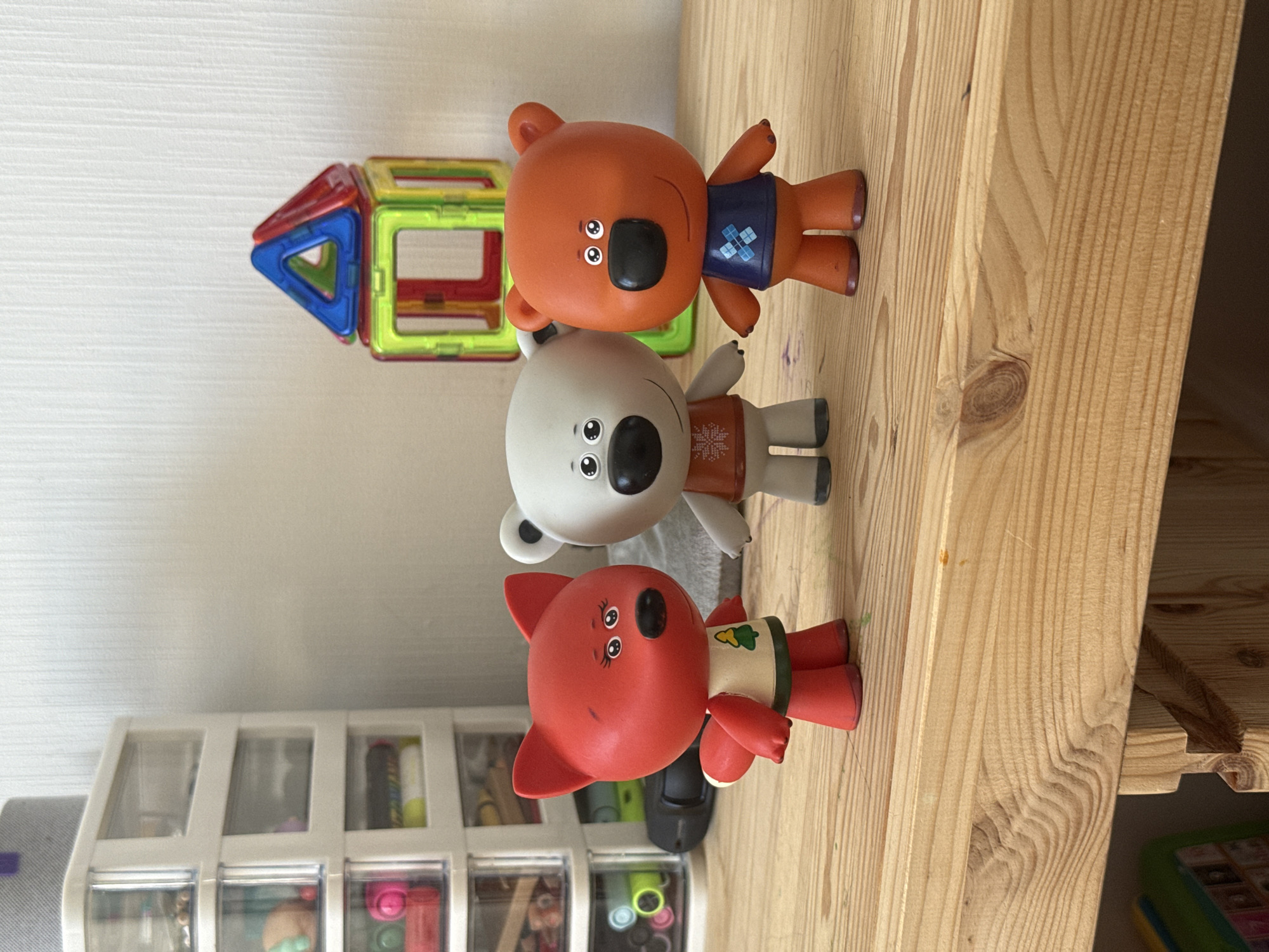 Three cartoon character figurines, a red fox and two bears, stand on a wooden surface. The red fox figurine, on the left, wears a white shirt with a green trim and a green tree graphic. The bear figurine in the center is white and grey, wearing an orange shirt with a white snowflake pattern. The bear figurine on the right is orange and wears a dark blue shirt with a light blue diamond pattern. Behind the figurines, a white wall is visible with a textured pattern. To the left of the figurines, a multi-drawer plastic organizer filled with various items is visible. To the right of the figurines, a colorful magnetic building toy set forms a structure resembling a house. The wooden surface appears to be part of a table or shelf, with visible wood grain and knots. The overall scene is indoors, likely in a domestic setting. The provided information does not contain location-specific details for Lyubertsy, Russia.