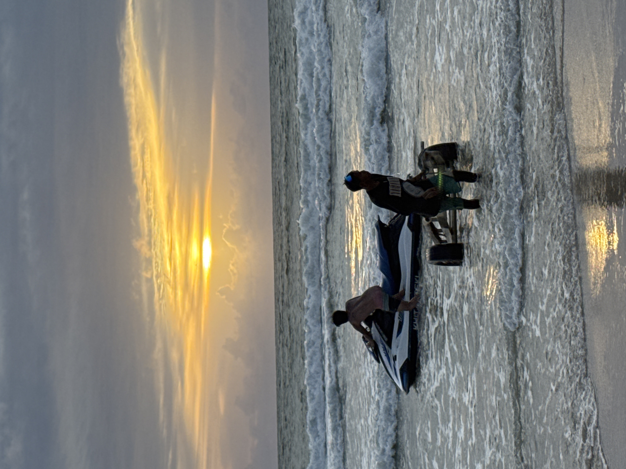 The image depicts a serene sunset scene at Patong Beach, Phuket, Thailand, with two individuals preparing a jet ski on the shore. The sky is a canvas of soft grey clouds tinged with the golden hues of the setting sun, which breaks through the clouds, casting a warm glow on the water. Gentle waves roll towards the sandy beach, creating a soothing rhythm.
In the foreground, a jet ski is partially submerged in the shallow water, its sleek white and blue body contrasting with the darker elements. One person, seen from the back, is mounting the jet ski, their physique suggesting a younger individual. They are wearing shorts. Beside them, another person, dressed in a black long-sleeved shirt and light-colored shorts, stands near a trailer-like structure with wheels, which appears to be used for transporting the jet ski. This individual is also partially in the water, their stance indicating they are assisting with the jet ski's movement or preparation. Their presence suggests a shared activity, perhaps readying the jet ski for an evening ride or for storage.
The overall atmosphere is tranquil, with the natural beauty of the sunset complementing the presence of human activity. The water is clear enough to see the sandy bottom in some areas, and the reflections of the sky and the jet ski are visible on the wet sand and the surface of the water. The scene captures a moment of peaceful interaction with the ocean and the vibrant colors of a tropical sunset.