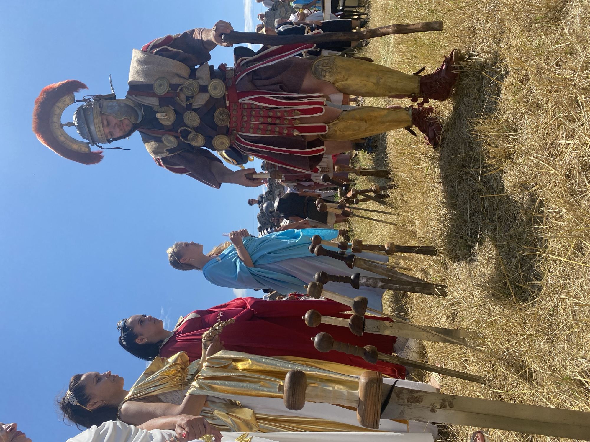 This image captures a historical reenactment or cultural event in the city of Gigen, Bulgaria, under a bright, clear blue sky. The foreground is dominated by dry, yellowish-brown grass and dirt, with strong shadows indicating intense sunlight.
In the center-right, a Caucasian male dressed as a Roman soldier stands prominently. He wears a detailed helmet (galea) with a large red plume (crista), segmented body armor (lorica segmentata style) adorned with multiple circular bronze medallions (phalerae) on leather straps, a brown tunic, and a red and white striped pteruges skirt. Golden greaves protect his shins, and he wears dark red, open-toed footwear. He holds a thick, gnarled wooden staff in his right hand, and a ring is visible on his right ring finger. His gaze is directed downwards, possibly at the swords in the ground.
To the left of the Roman soldier, a line of three women in ancient-style costumes stands. The leftmost woman is partially visible, wearing a white toga-like garment. Next to her is a dark-skinned woman with dark hair and a golden headpiece, adorned in a flowing golden toga-like dress. Her expression is serious, and she looks to her right. She holds a small golden, ornate object in her left hand. Beside her, a dark-skinned woman with dark hair tied back wears a red dress and a small tiara, also looking to her right with a serious expression. Further to the right, a fair-skinned woman with blonde hair and a golden headpiece is dressed in a light blue toga-like gown. She holds a small white object in her right hand.
In the immediate foreground, between the women and the soldier, approximately twelve wooden swords with varying hilt designs are stuck vertically into the dry ground.
In the blurred background, behind the Roman soldier, indistinct figures of other people and some light-colored stone structures or ruins are visible, suggesting an active event or a historical site.
