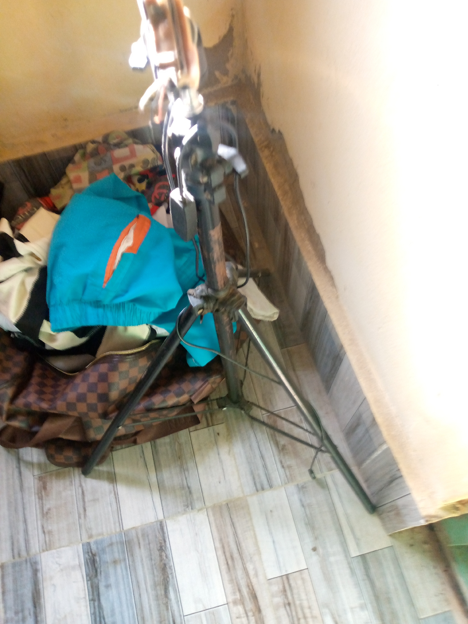 In Jalingo, Nigeria, a scene unfolds within a room featuring walls with peeling paint and a floor covered in wood-patterned tiles. In the foreground, a large, dark brown checkered patterned bag is partially visible, situated on the tiled floor. Above the bag, a pile of clothes is present, including a vibrant turquoise jacket with an orange accent. Dominating the center of the image is a metal tripod stand with visible wires and a black box attached to it. A portion of a light-colored wall and a beige ceiling are also captured in the frame. The lighting in the room appears to be from an overhead source, creating strong highlights and shadows.
