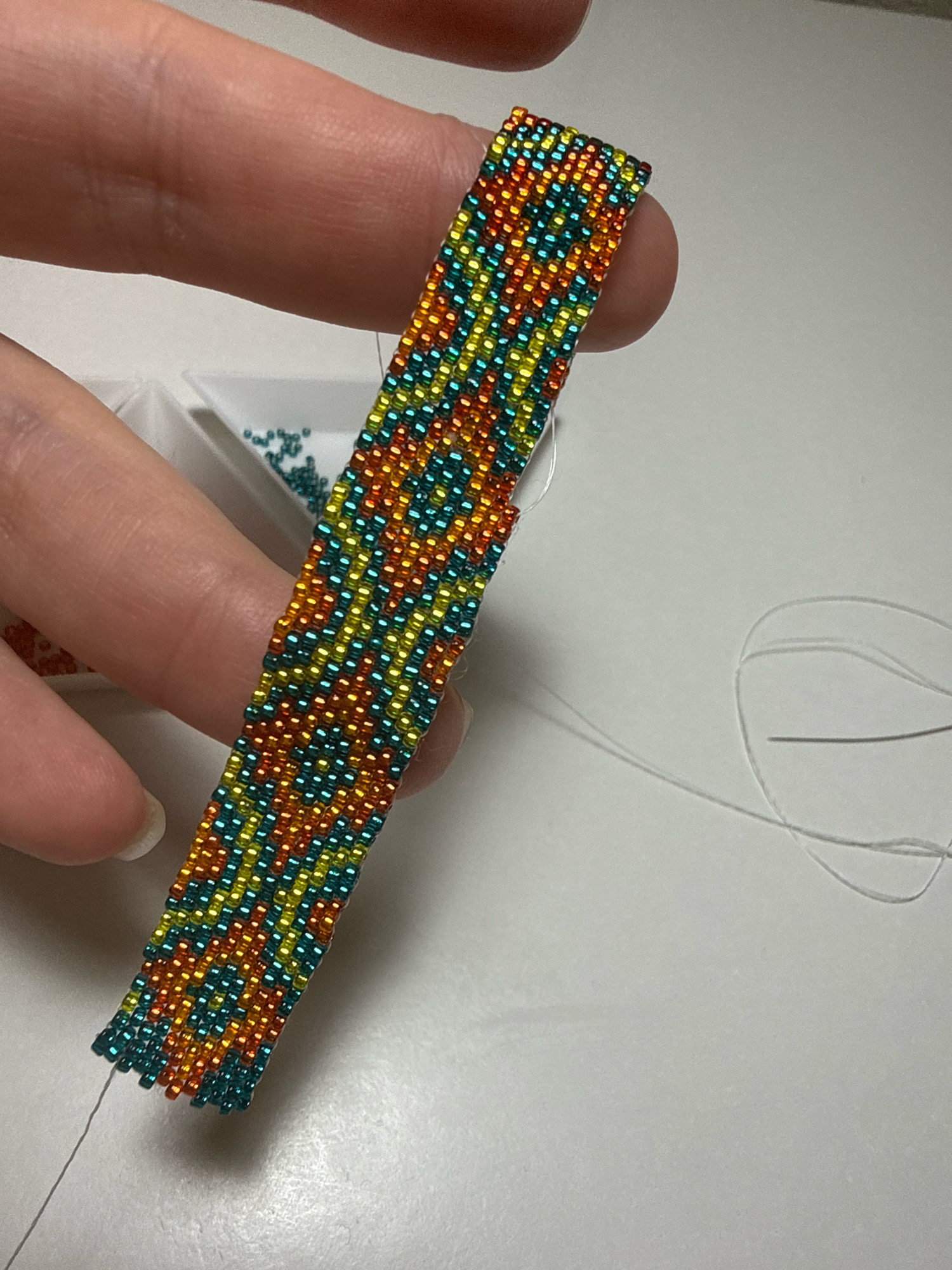An individual's fingers hold a beaded bracelet that is approximately two inches wide and four inches long. The bracelet is made of small, iridescent seed beads in alternating rows of teal, yellow, and orange. The beads are arranged in a pattern that creates diagonal stripes. The bracelet is being held over a small white plastic tray containing a scattering of teal seed beads. A thin white thread extends from the bottom edge of the bracelet and disappears out of frame to the right, where it forms a loose loop. The background is a plain, light-colored surface, likely a table. The image is a close-up shot, with the focus on the bracelet and the hands holding it. There is no discernible connection to Kazan, Russia, in this image.