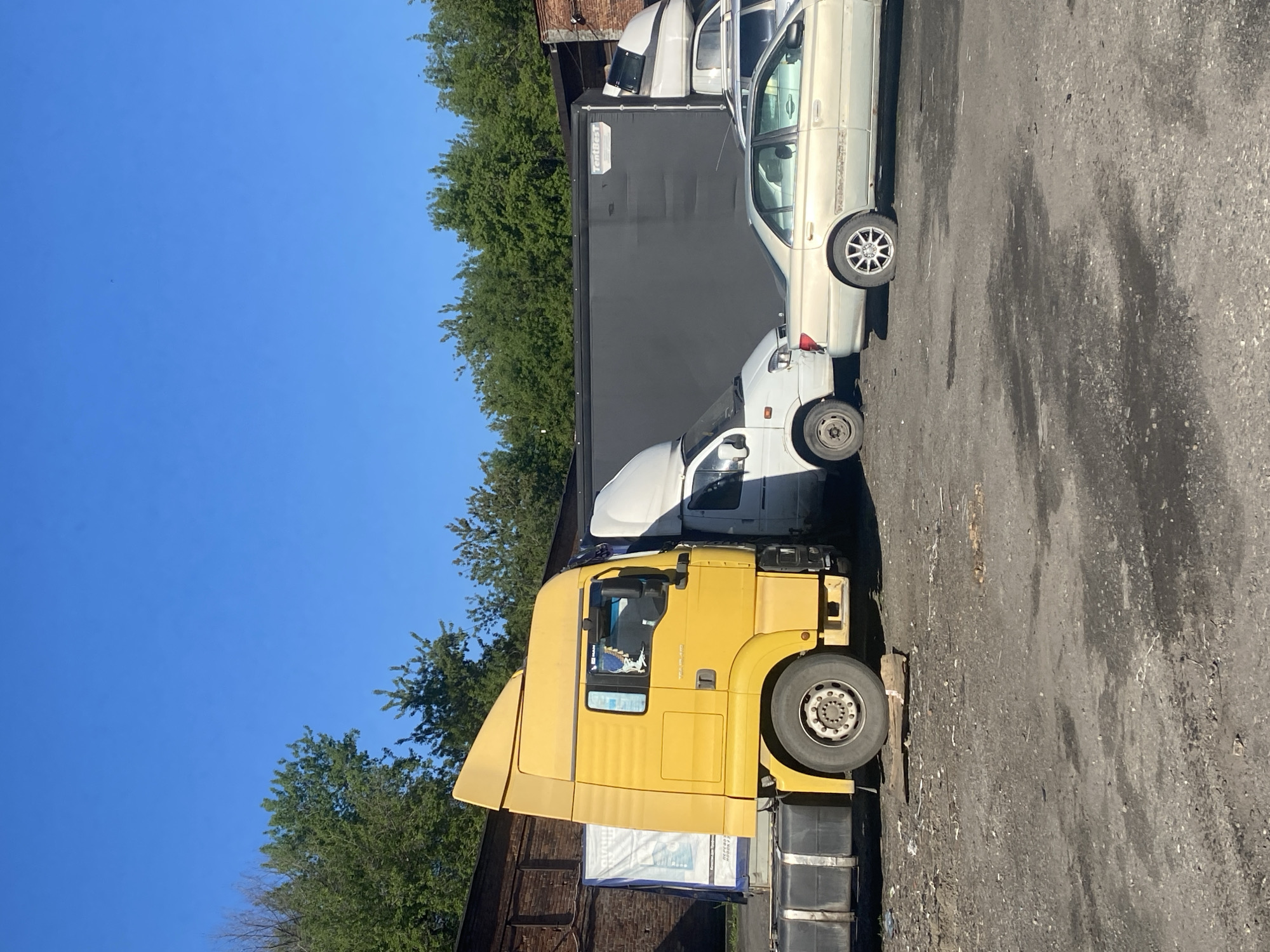 The image is presented in a rotated orientation, appearing as if taken in portrait mode and viewed horizontally. When viewed correctly, it depicts an outdoor parking or industrial area in Novokuznetsk, Russia, under a clear, bright blue sky.
The scene is illuminated by strong daylight, with sunlight casting subtle shadows. Several vehicles are parked on a dark, uneven asphalt or paved surface. On the left side of the frame, a prominent yellow semi-truck is visible, featuring the "MAN" logo on its cab. This truck is hitched to a long, black semi-trailer, which has the white text "TentBest" printed on its side.
Towards the center and right of the frame, a white delivery van is parked behind the truck. Further to the right, a light-colored sedan, possibly beige or silver, is parked. Another white vehicle, likely a truck cab, is partially visible behind the sedan.
In the background, a dense line of lush green deciduous trees creates a natural boundary. Portions of a dark building or industrial structure can be seen through and above the foliage. There are no people visible in the image.