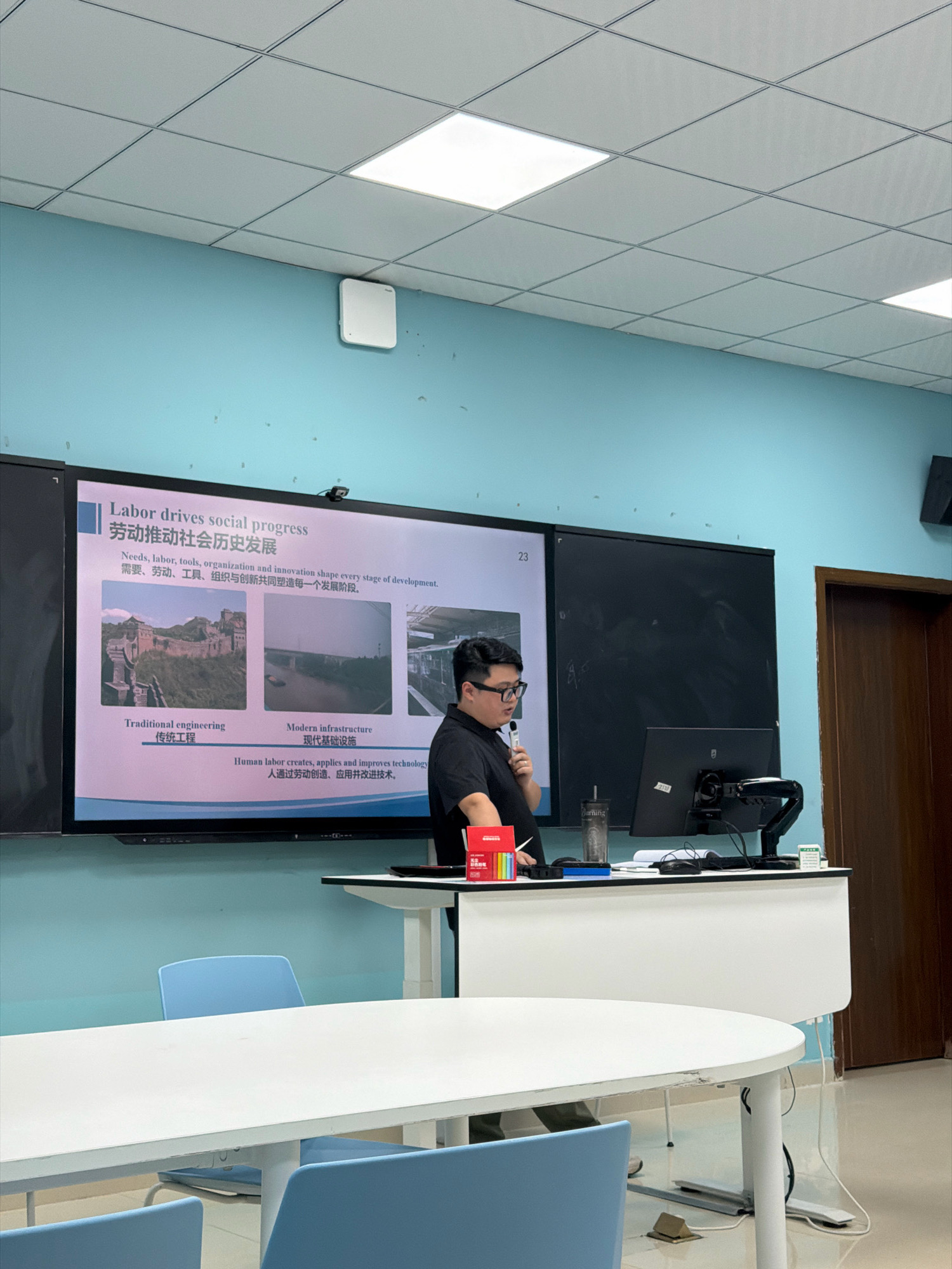In a classroom, a male lecturer stands at a podium facing a large projection screen. The screen displays a presentation titled "Labor drives social progress" in English and Chinese, with images of traditional engineering (the Great Wall) and modern infrastructure. The lecturer, wearing glasses and a black polo shirt, holds a microphone and appears to be speaking. The podium is equipped with a computer monitor, a document camera, and other presentation equipment. The room has blue walls and a white ceiling with fluorescent lights. A whiteboard is visible behind the lecturer. Several blue chairs and white tables are arranged in the classroom, suggesting it is a learning environment. The scene appears to be set in an educational institution, potentially a university or vocational school. However, there is no information in the image to determine the specific location in Jiangjin, China.