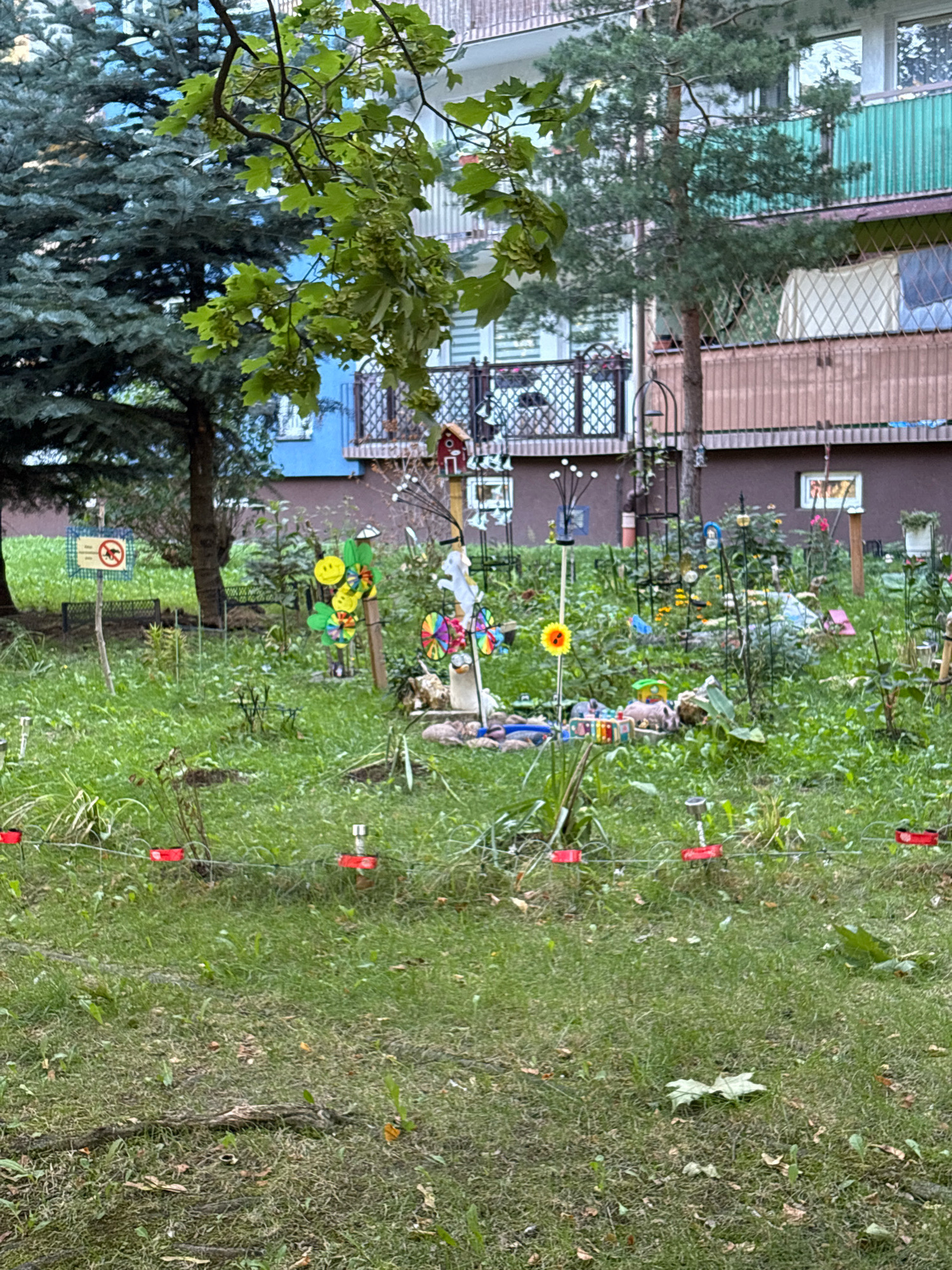 The image depicts a small, cluttered garden area in front of a multi-story residential building. The garden is densely planted with various types of vegetation, including grass, small trees, and flowering plants. Many decorative objects are scattered throughout the garden, including colorful pinwheels, small figurines, and what appears to be a birdhouse. A wire fence with red markers runs across the foreground. In the background, the building has several balconies with railings and windows. The scene is brightly lit, suggesting it is daytime. The specific location in Wrocław, Poland, is not discernible from the visual information provided.