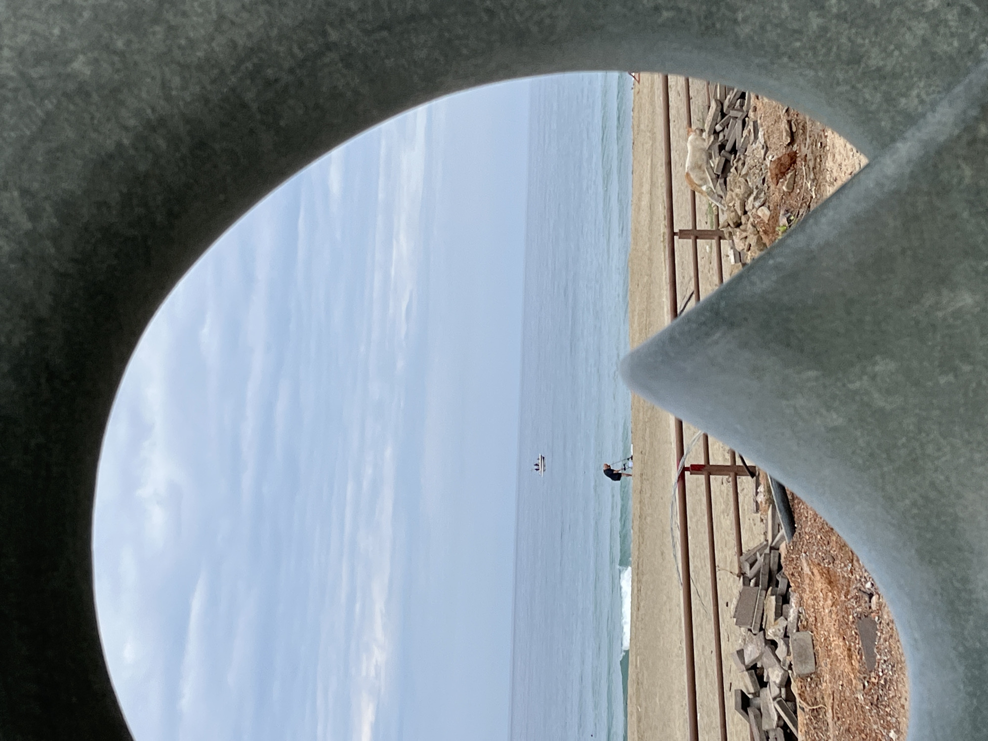 The image, seemingly taken from a viewpoint in Alanya, Turkey, frames a coastal scene through a circular aperture of a gray, concrete structure. The horizon line is nearly centered within the circular frame, dividing the sky and sea. Above, wisps of clouds streak the light blue sky. Below, the sea mirrors the sky's color, with gentle waves breaking on a sandy beach.
A person stands at the shoreline, to the right of a sailboat in the sea. Adjacent to the beach, a section of the concrete structure is visible, interspersed with pieces of brick and dirt. A light-colored dog stands atop the embankment, appearing to look towards the ocean.