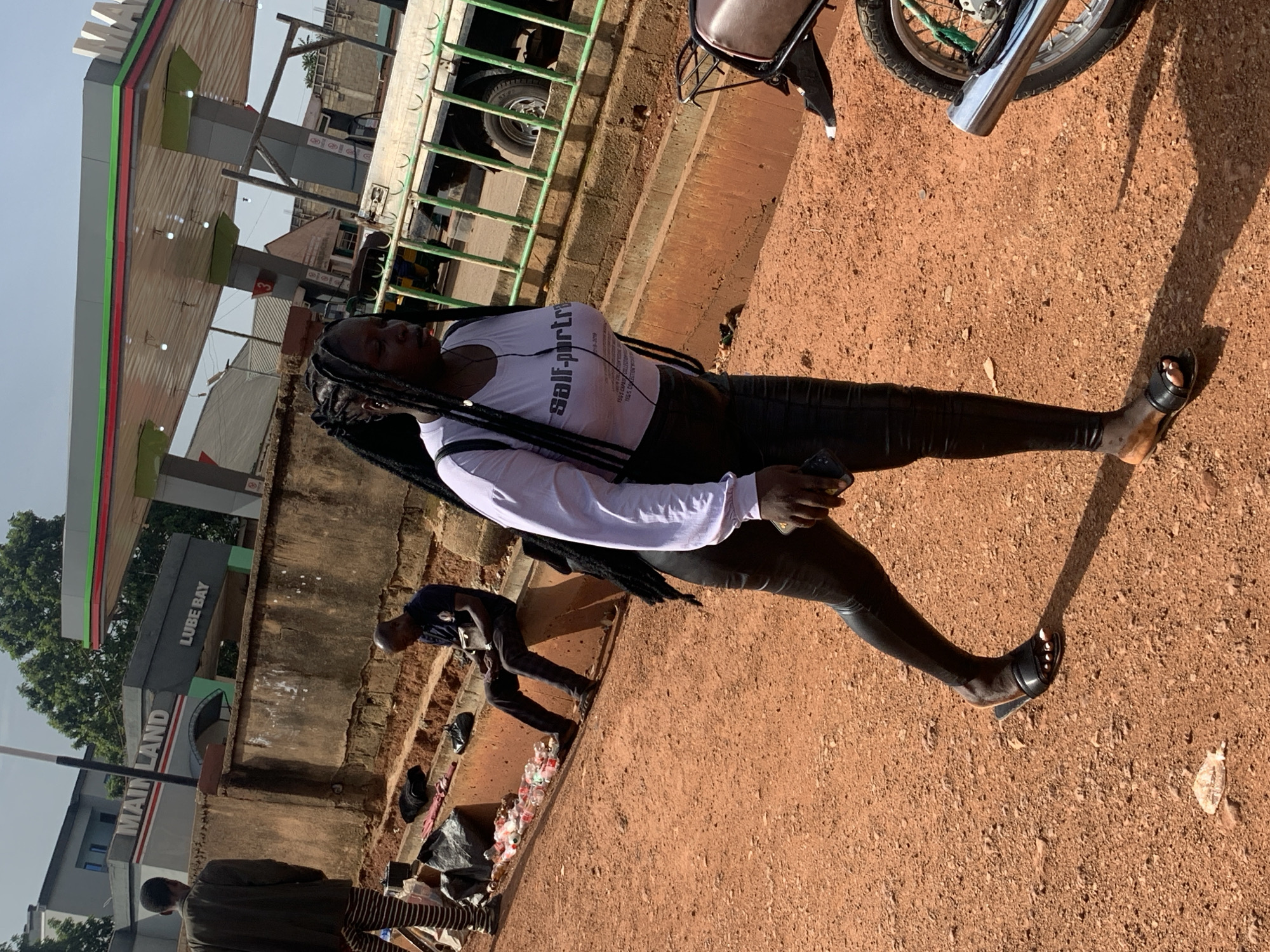 A young Black woman with long braided hair is walking in the foreground on a dusty, orange-red dirt path. She is wearing a white long-sleeved t-shirt with the words "Self-portrait" printed in black on the front, black tight-fitting pants, and black open-toed sandals. She is carrying a black bag over her shoulder and holding a smartphone in her right hand.
In the middle ground, a man wearing a dark blue polo shirt and dark pants is sitting on a concrete ledge, looking down at his hands. Several plastic water bottles are lined up on the ledge in front of him. Behind him, a row of bags and clothing items are spread out.
In the background, a gas station with a red, green, and white canopy is visible. Signs on the gas station read "MAIN LAND" and "LUBE BAY". A large truck with a green railing is parked under the canopy. The environment appears to be an urban or semi-urban area in Nigeria. The overall scene depicts a sunny day in a bustling outdoor environment.