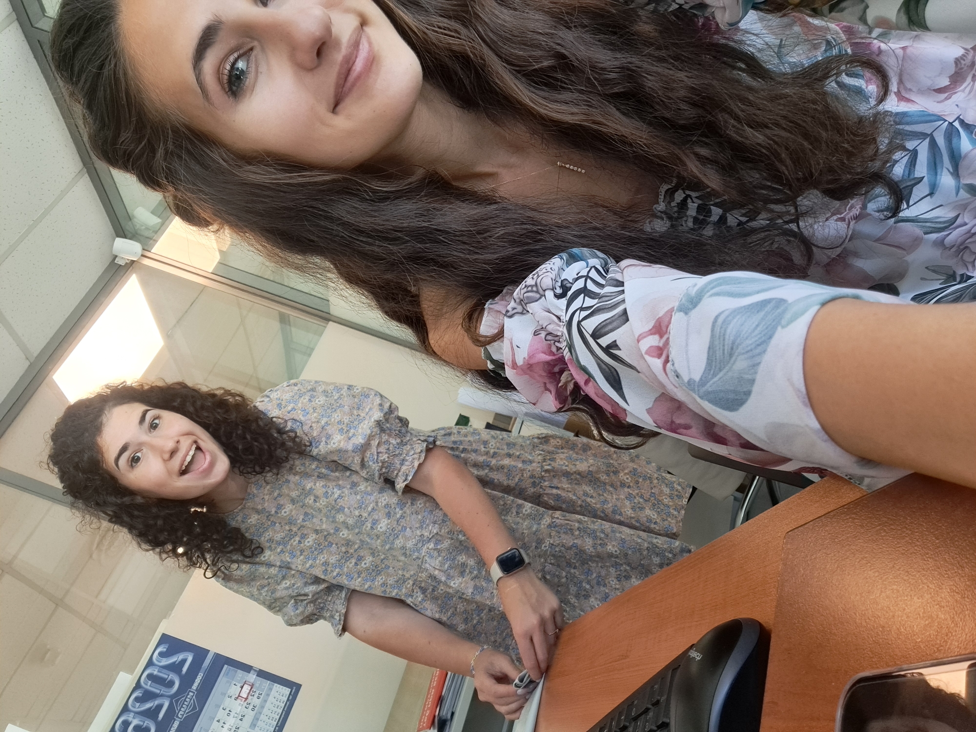 In a brightly lit indoor environment, possibly an office in Sofia, Bulgaria, two young women are pictured. The woman in the foreground, with long, dark, wavy hair, is positioned to the right of the frame, facing slightly towards the camera. She has a warm smile and is wearing a floral-patterned top with large pink and white flowers. A delicate silver necklace with a pendant hangs around her neck. Her hair is partially pulled back, framing her face.
To the left, another woman with curly, dark brown hair is smiling broadly with her mouth open, looking directly at the camera. She is wearing a light-colored dress with a blue and beige floral pattern. Her dress has short, puffed sleeves. She is wearing a smartwatch on her left wrist and a ring on her left hand. Her hands are positioned as if she is holding or examining a piece of paper.
The background reveals office-like elements. A calendar with the month "SASOS" visible is mounted on the wall to the left. The environment appears to be a modern office space with glass partitions and fluorescent lighting. A desk with a black computer keyboard and mouse is in the lower part of the image, suggesting a work setting. The overall scene captures a candid moment between two individuals in a professional or semi-professional setting.