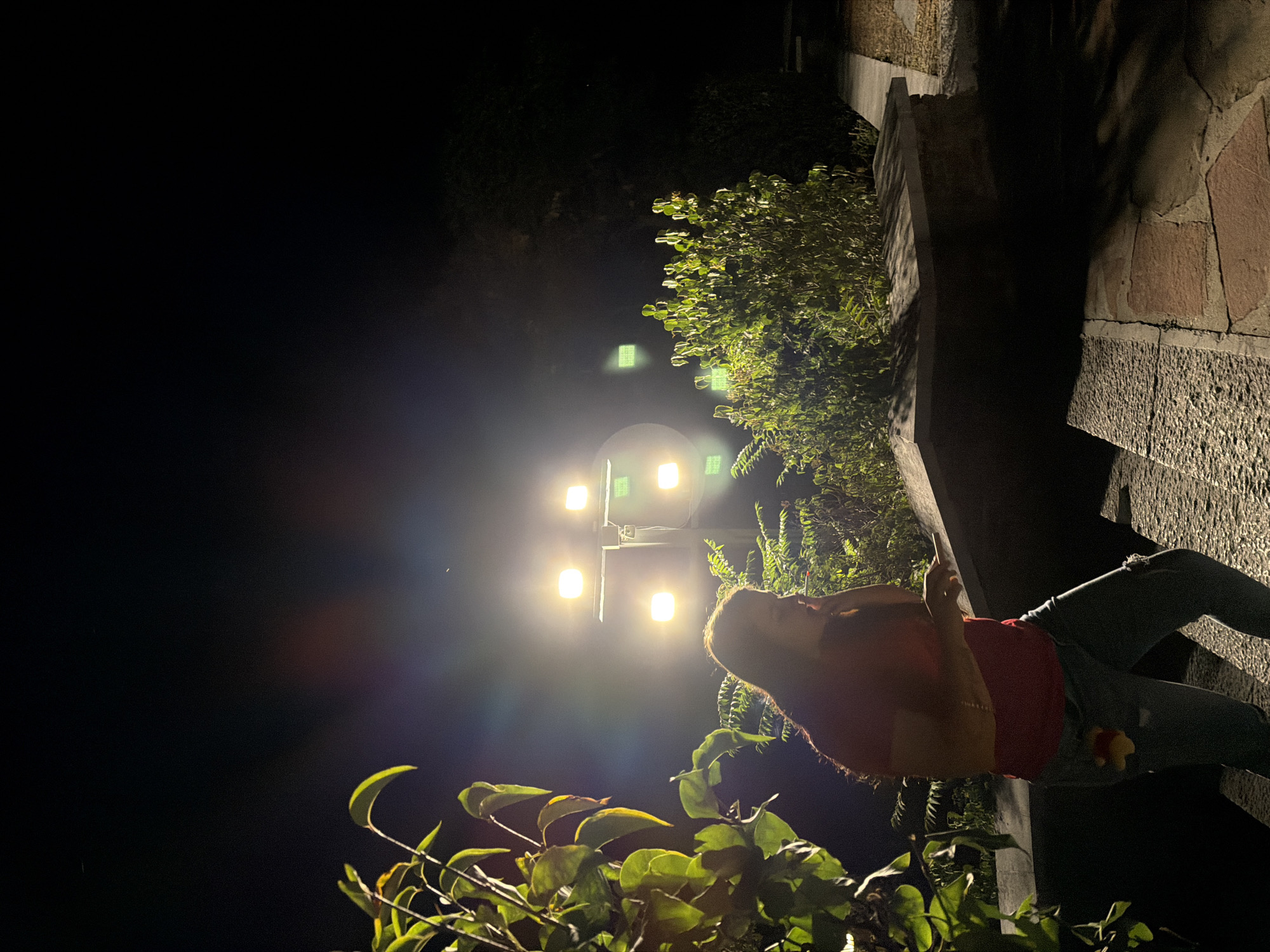 A person stands on stone steps at night, illuminated by multiple bright floodlights mounted on a pole. The person, wearing a red shirt and ripped jeans, holds a smartphone in their right hand and appears to be speaking into it or gesturing towards it. A yellow charm is attached to their jeans. Lush green foliage frames the scene on the left, with more greenery visible behind the person and to the right. The ground is paved with stone slabs and bricks. The sky is dark, with a subtle lens flare or light pollution visible. This scene appears to be located in the vicinity of the park near the Lovech fortress in Lovech, Bulgaria.