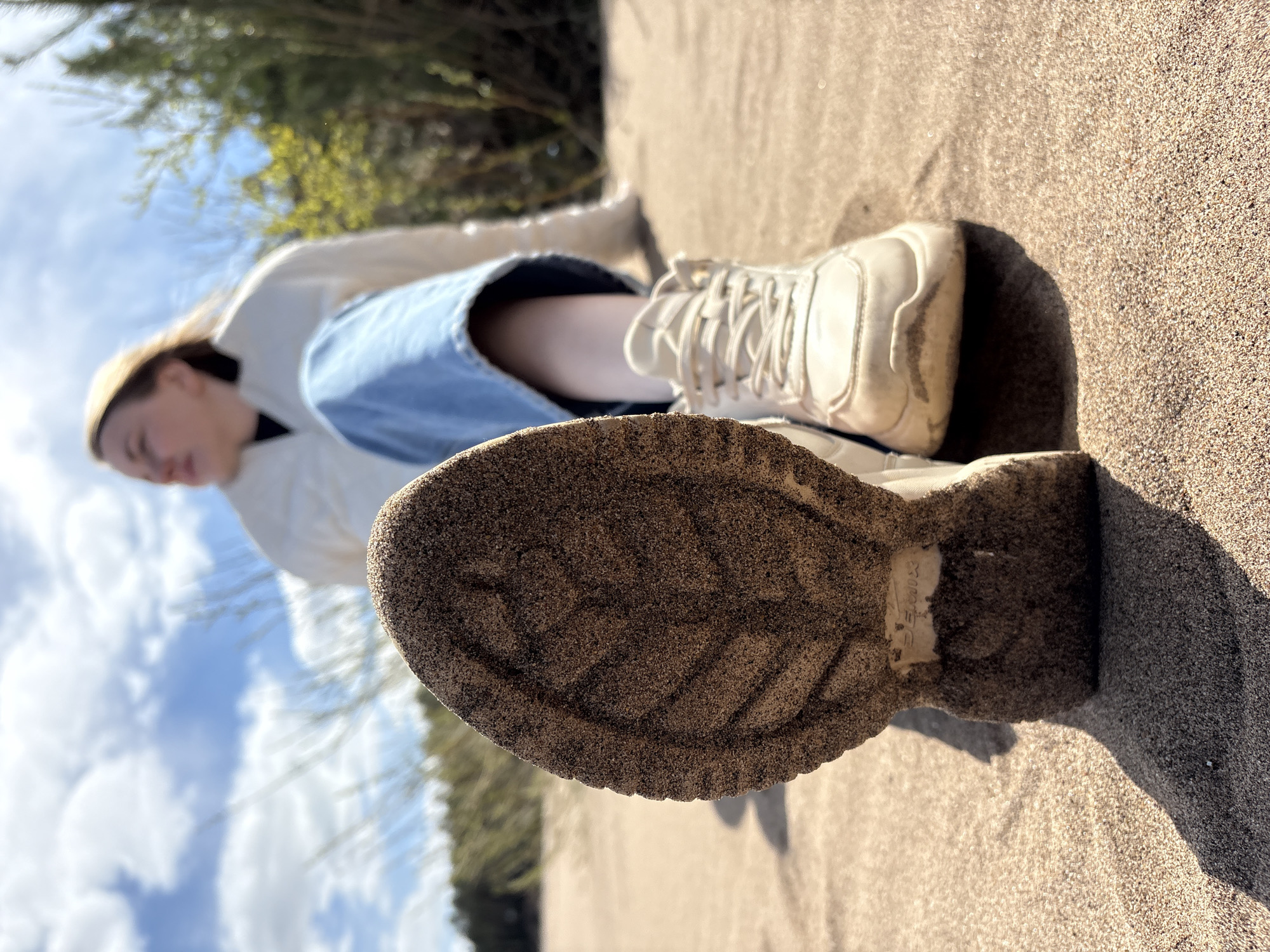 A horizontal, close-up shot shows a person lying on a sandy surface, likely a beach. The person is wearing blue denim shorts, a white shirt, and beige shoes. The sole of one shoe is prominently in the foreground, covered in sand. The sand is coarse and light brown. The background shows a blurred view of trees and a blue sky. Based on the prompt, the location is Chaikovsky, Russia.